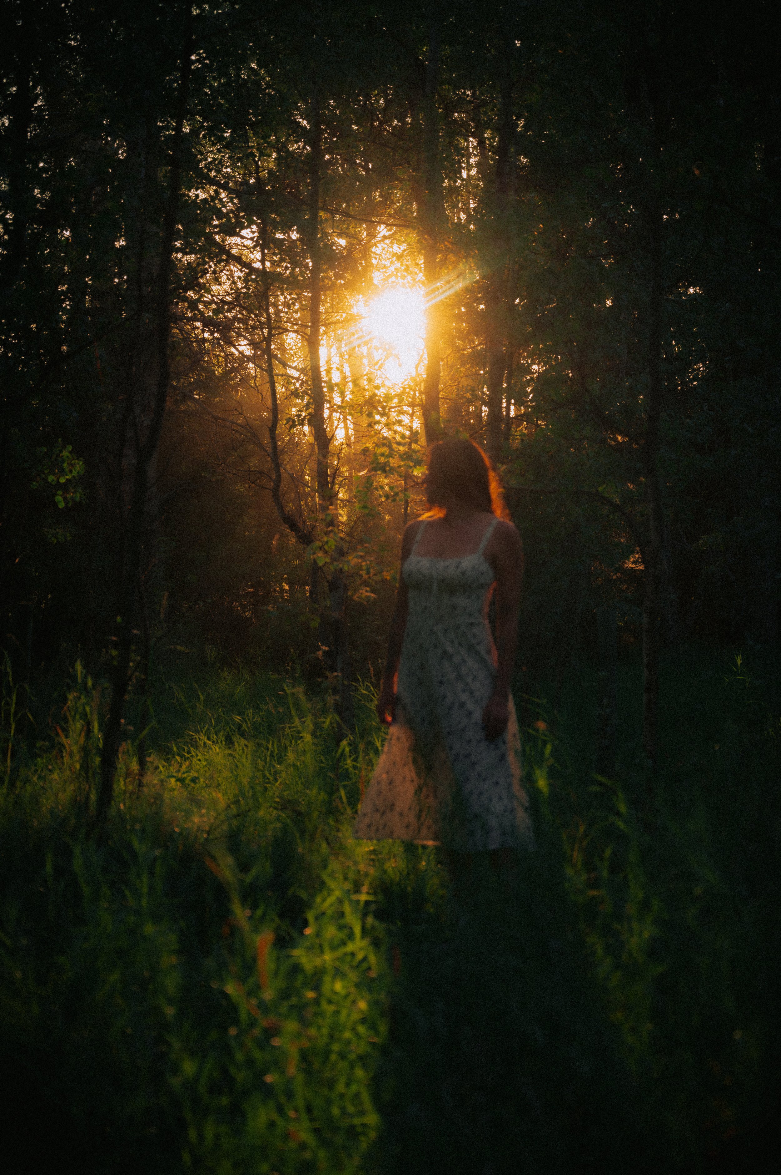 A woman walking in a forest during sunset with sunlight filtering through trees.