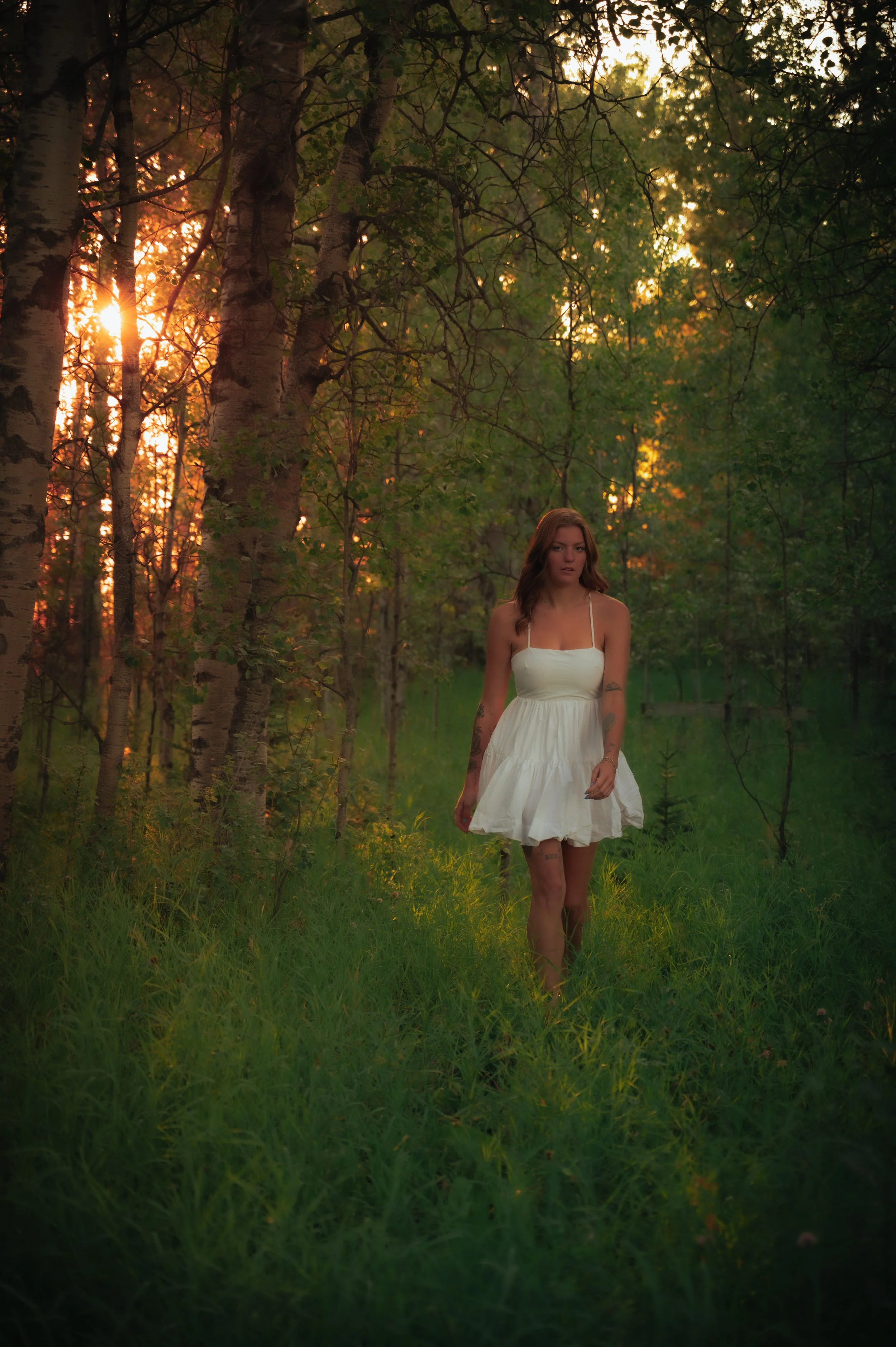 A young woman in a white dress walking through a green forest during sunset.