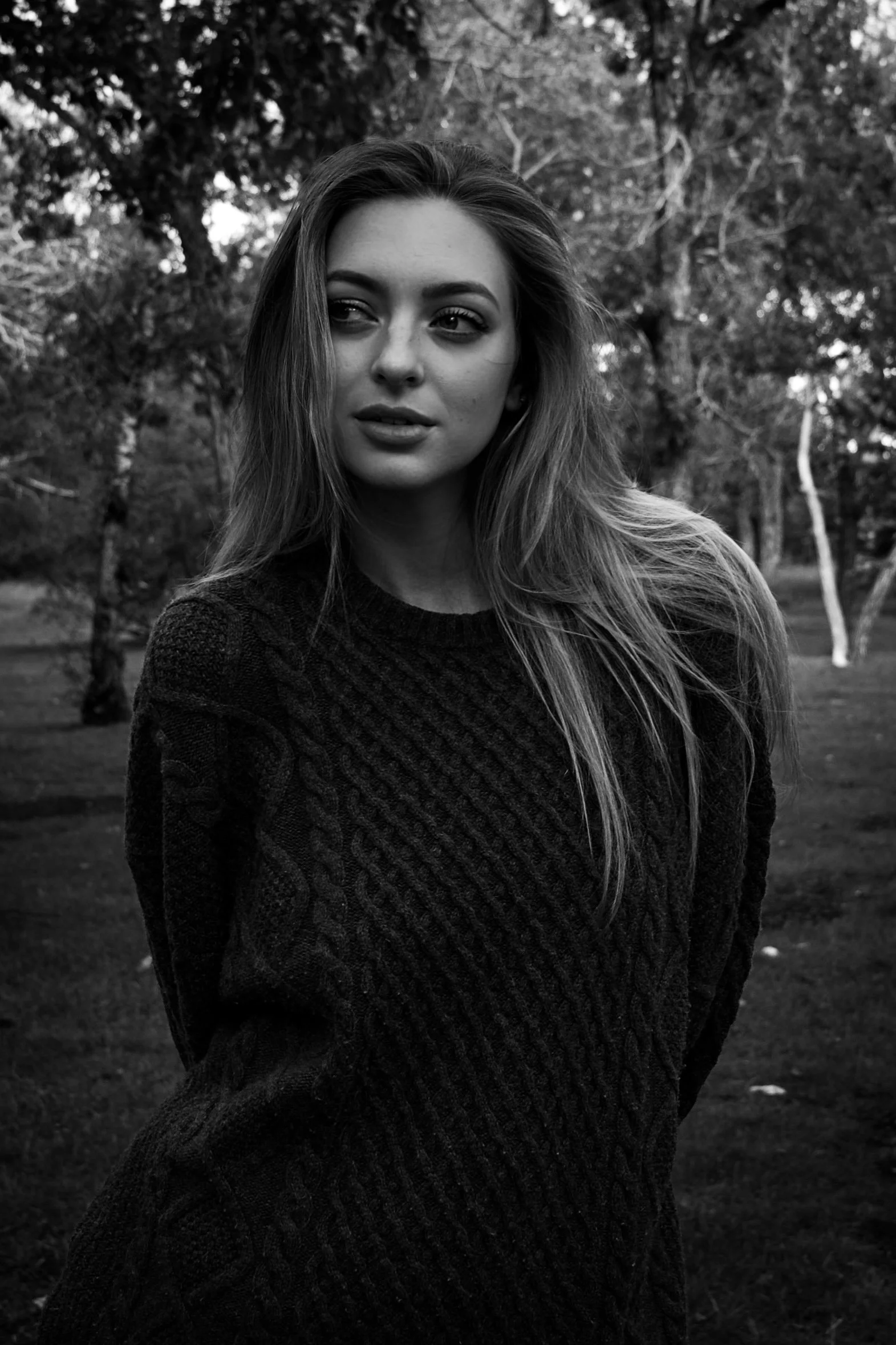 A black and white photo of a woman with long hair, outdoors with trees in the background.