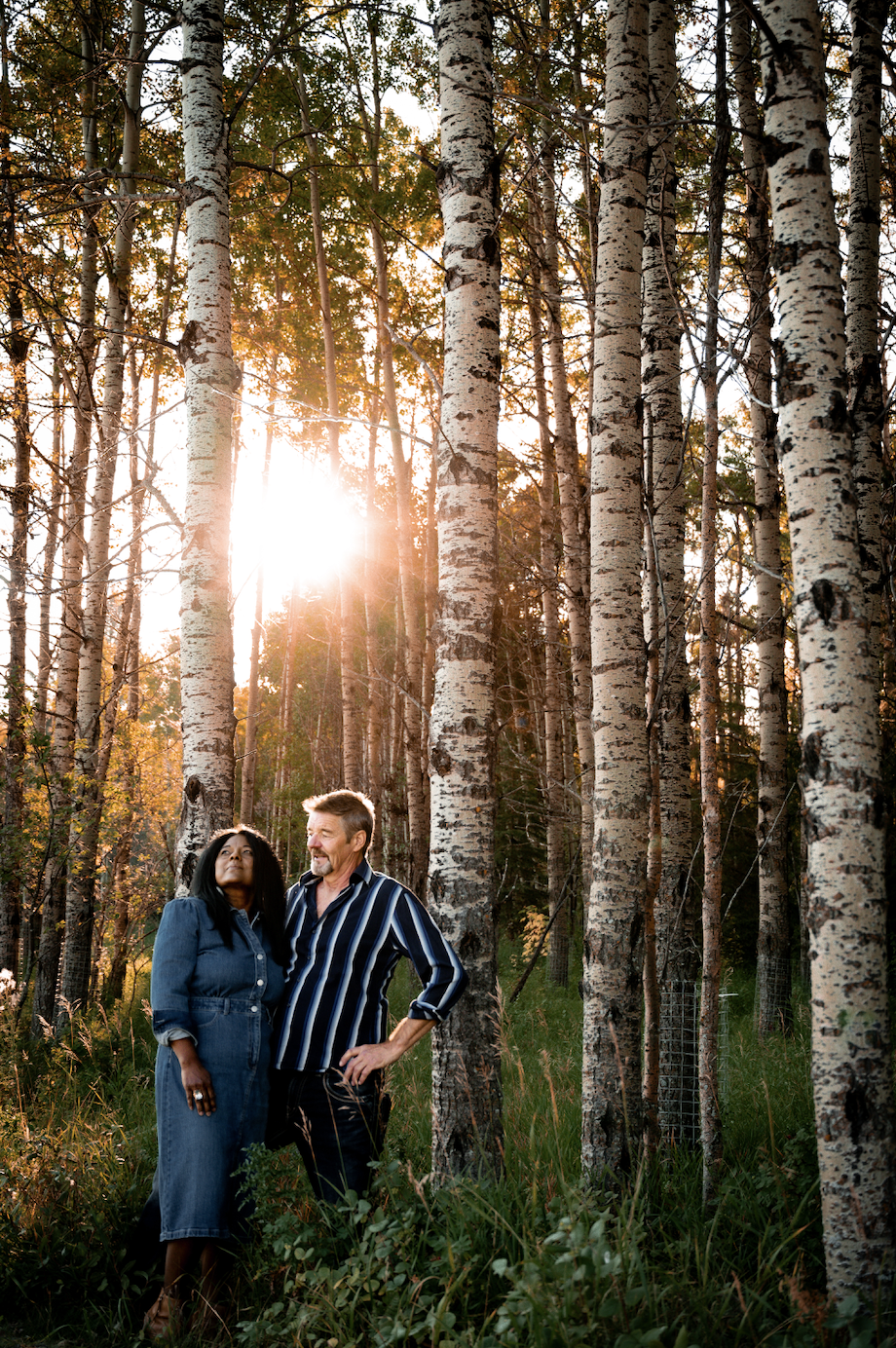 A couple standing in a birch forest during sunset, with sunlight shining through the trees, the woman is dressed in a denim jumpsuit, and the man in a striped shirt.