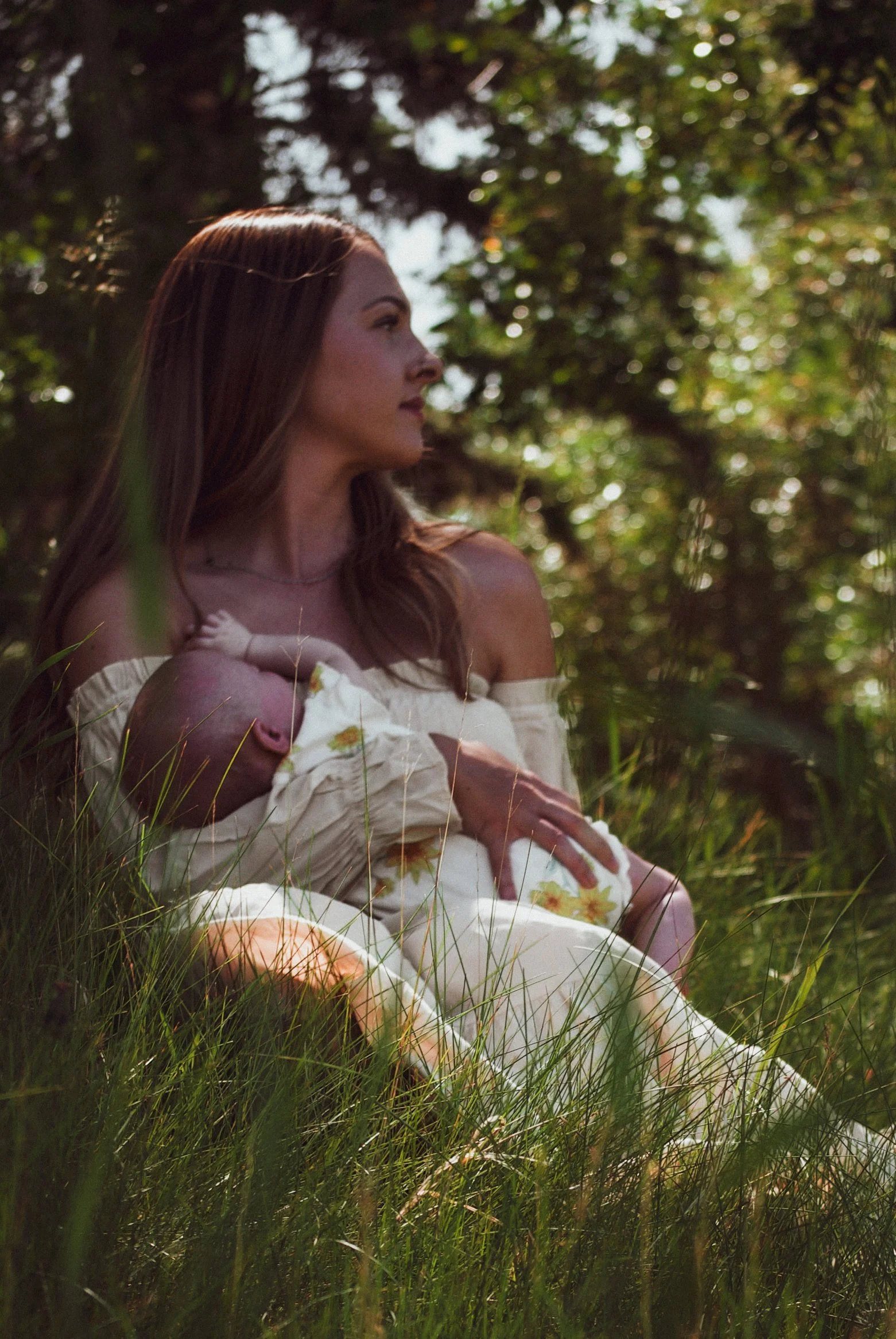 Woman with long red hair sitting in a grassy area, breastfeeding a baby wrapped in a white blanket with sunflower patterns, surrounded by trees.