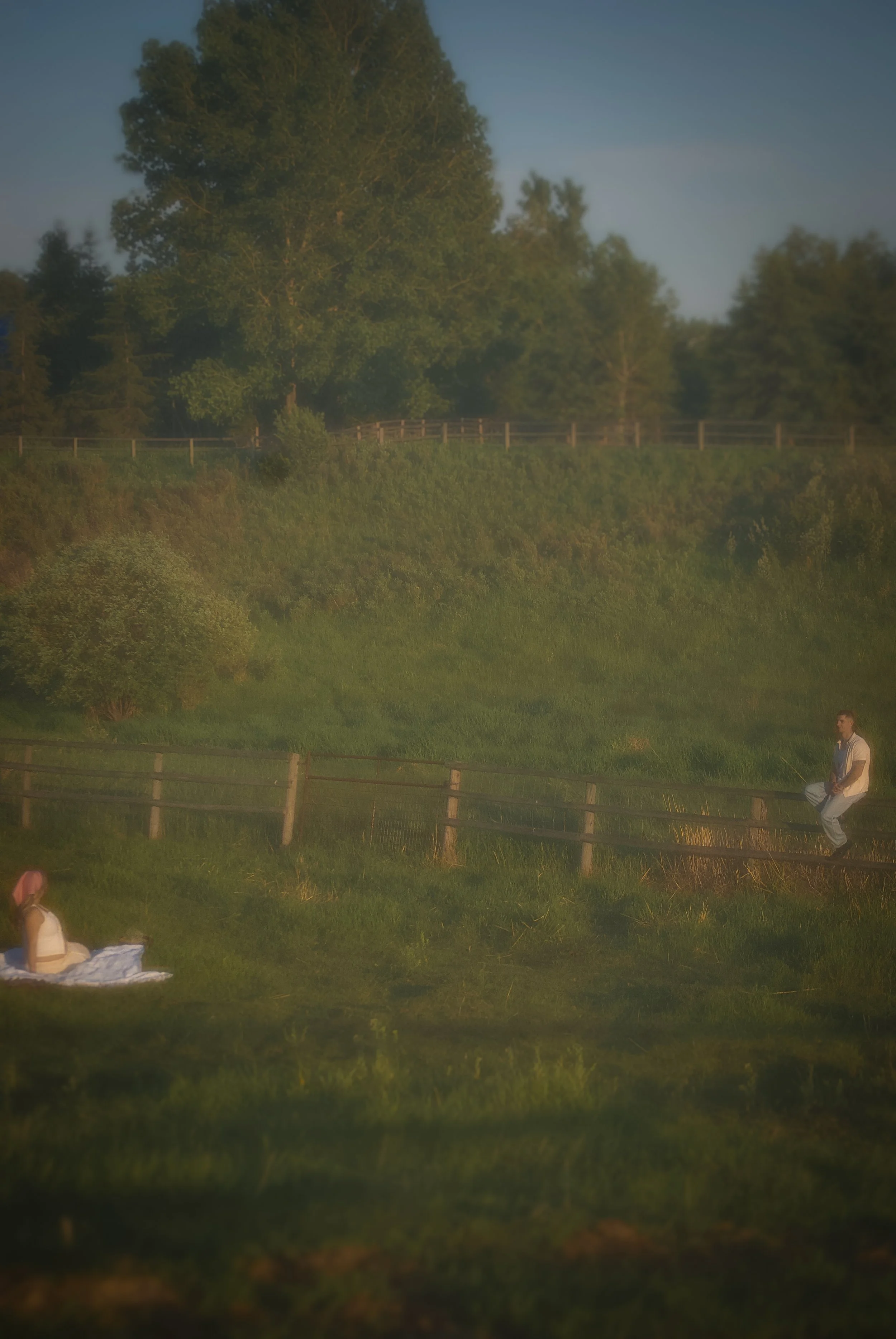 A person with dark hair sitting on a picnic blanket in a grassy field, wearing a pink hat and cream-colored top. Another person with dark hair and a beard is sitting on a wooden fence, playing a drum. The background features a lush, green hillside wi