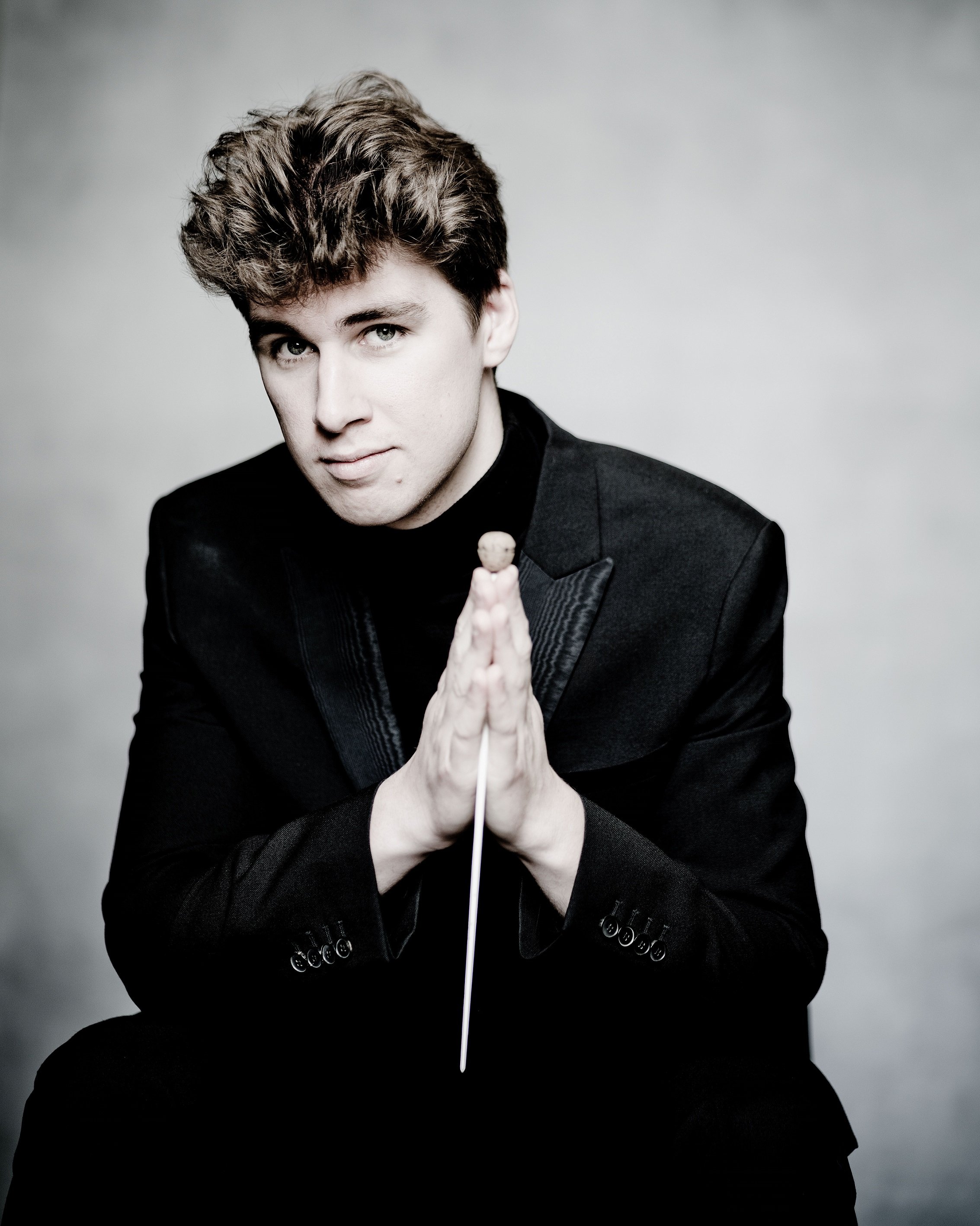 Swann van Rechem, a young man with brown hair and pale skin wearing a black suit, sitting with his hands pressed together in a prayer-like gesture, holding his baton. He is looking confidently into the camera against a gray background.