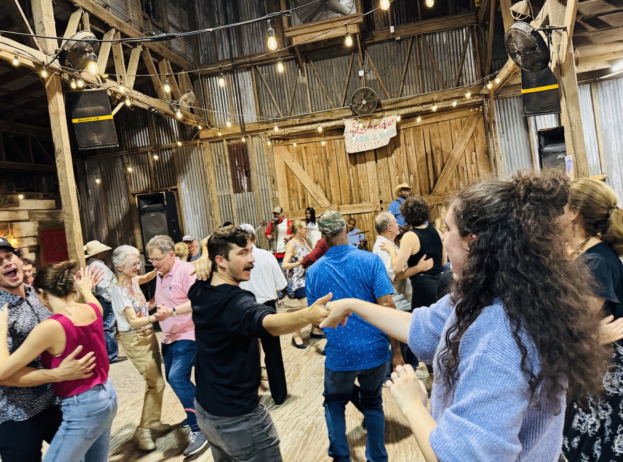 People dancing and enjoying music inside a rustic barn with string lights, speakers, and a banner that reads 'Lakeview Park & Beach.'