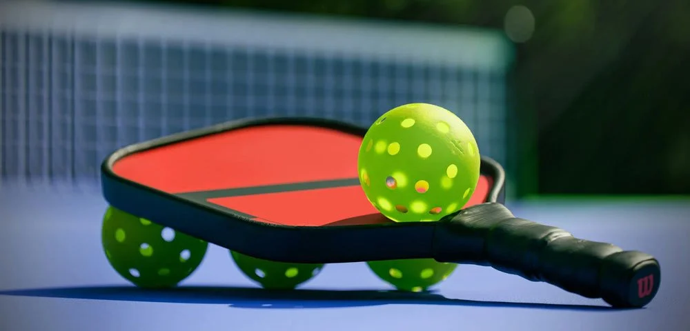 A pickleball paddle with three balls, all of which are bright green, lying on a pickleball court.