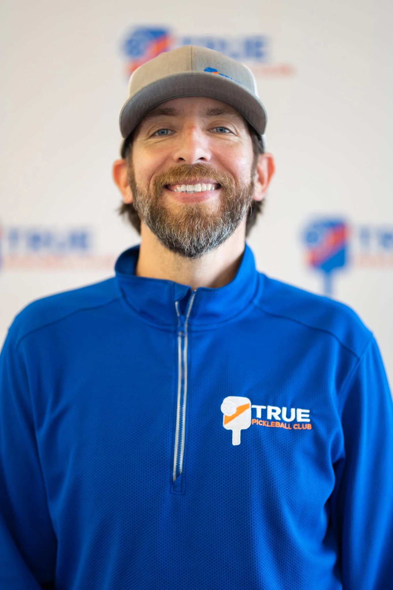 A man with a beard smiling, wearing a gray baseball cap and a blue jacket with a 'True Pickleball Club' logo, standing in front of a blurred background with similar logos.