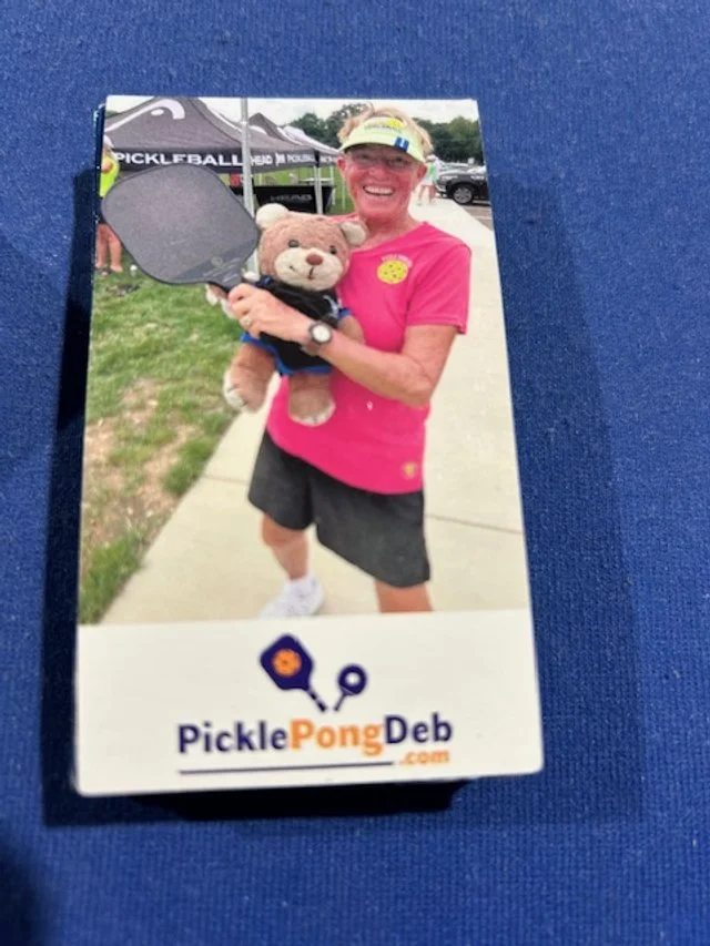 A woman smiling and holding a teddy bear and pickleball paddle at an outdoor event, with a sign reading "PicklePongDeb.com" at the bottom of the photo.