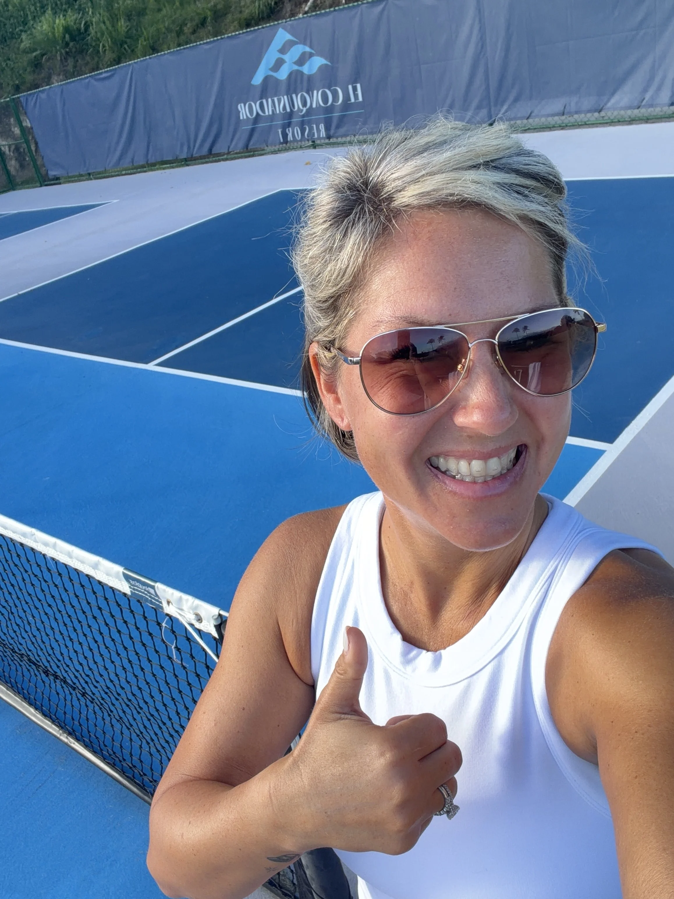 Woman smiling, wearing sunglasses, white tank top, giving a thumbs-up, next to a blue pickleball court with a net, outdoor setting, clear weather.