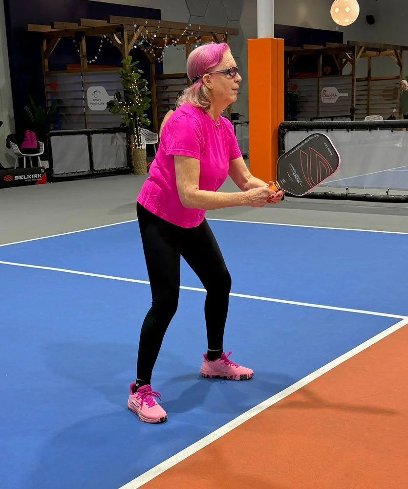 A woman with pink hair, wearing glasses, a pink t-shirt, black leggings, and pink athletic shoes, is standing on a blue and orange indoor pickleball court, holding a black racket with orange accents in a ready position.