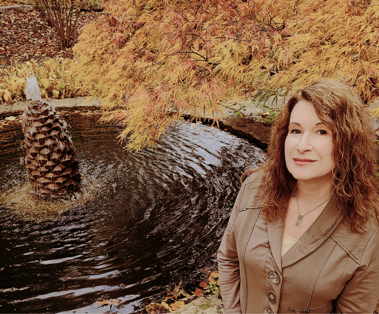 Picture of Trilogy Marketing Solutions Founder. A woman with curly brown hair wearing a tan jacket and a necklace, standing near a small pond with a pine cone fountain, surrounded by colorful autumn leaves and Japanese Maple tree.