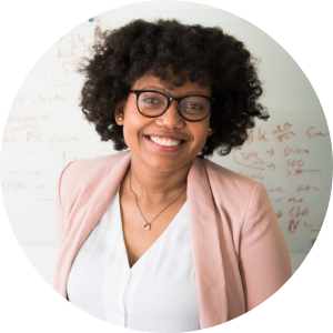 Smiling woman with curly hair and glasses, wearing a light pink blazer and white shirt, standing in front of a whiteboard with writing.