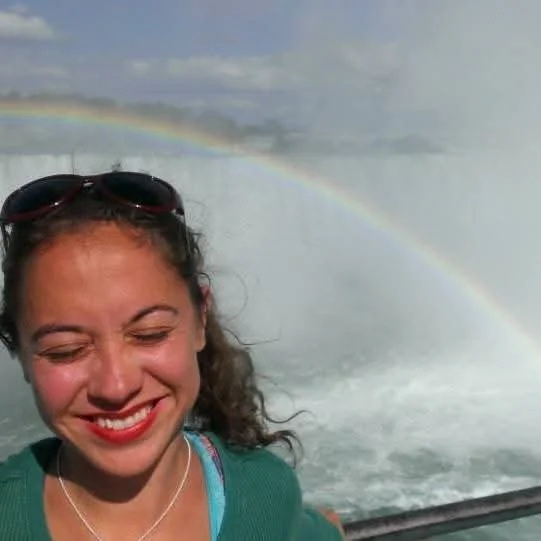A woman smiling in front of a waterfall with a rainbow in the background.