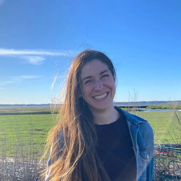 A young woman with long brown hair smiling outdoors in a grassy landscape with a clear blue sky.