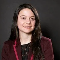 Portrait of a young woman with long dark hair, wearing a burgundy blazer and black top, smiling against a dark background.