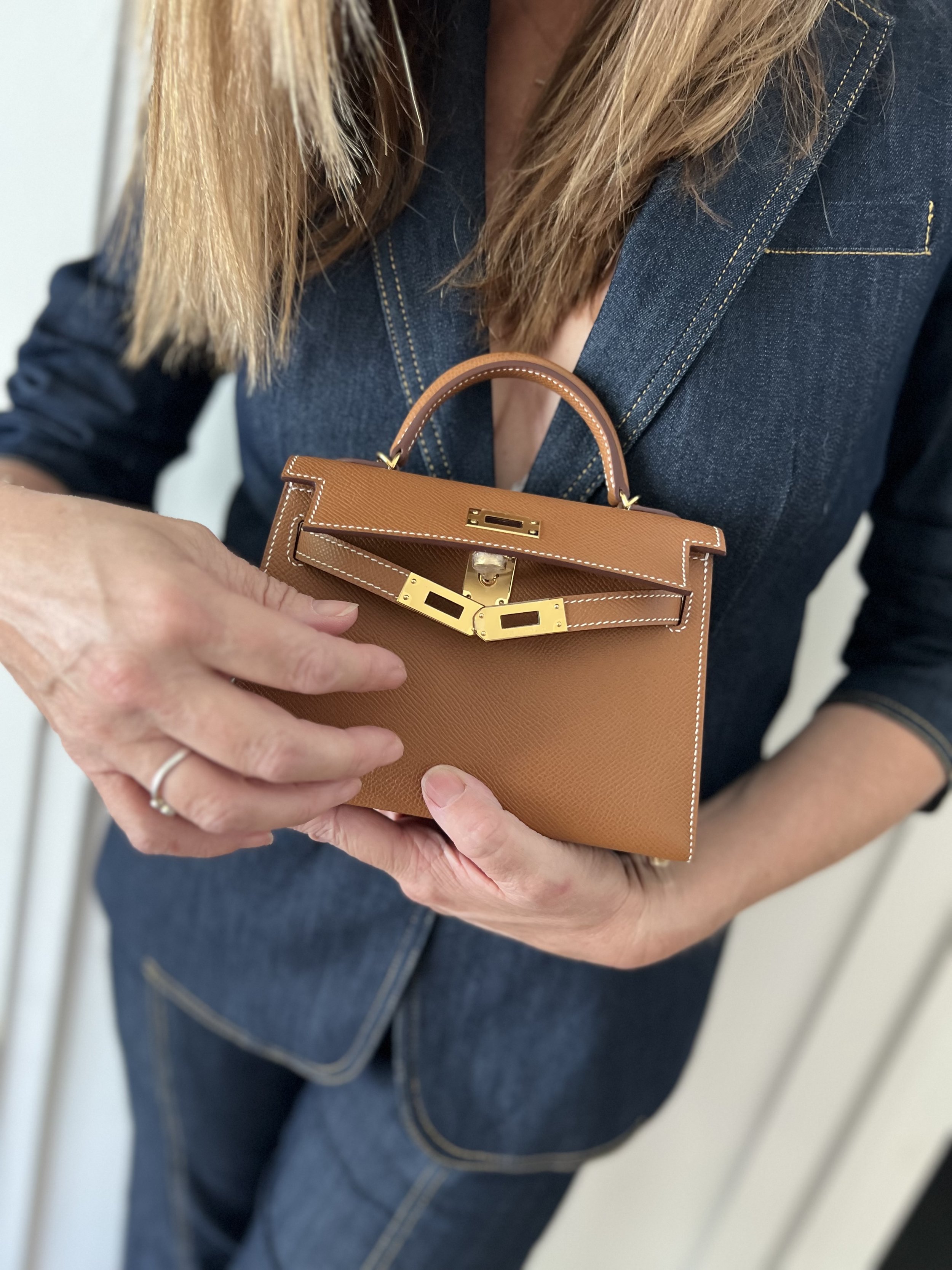 A woman in a dark denim suit holding a small tan handbag with gold hardware.