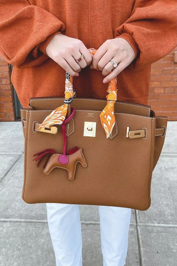 Person wearing an orange sweater holding a brown handbag with a decorative horse keychain, on a sidewalk with a brick wall in the background.