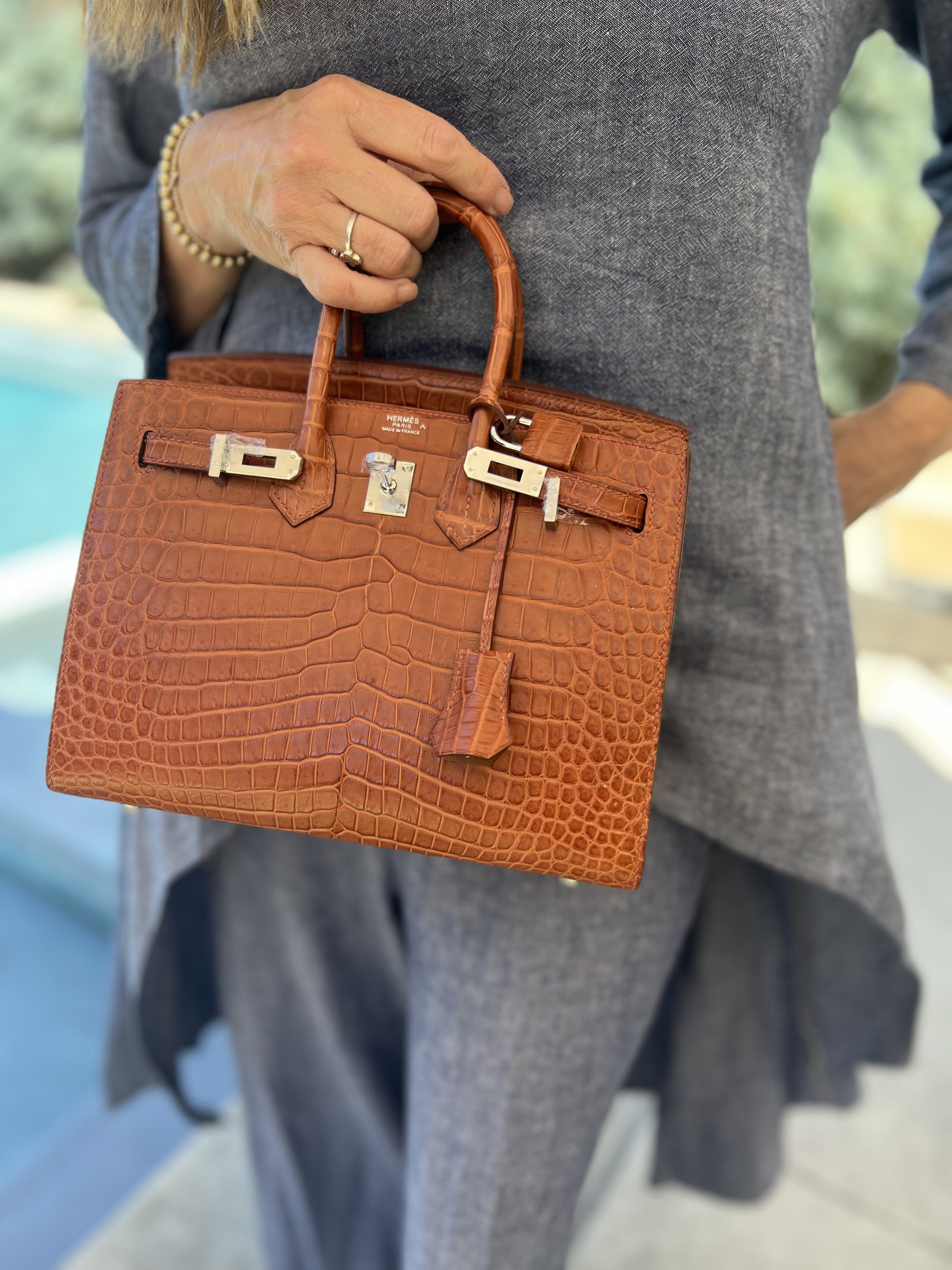Person holding a brown crocodile leather Hermès Birkin handbag with gold hardware, outdoors.