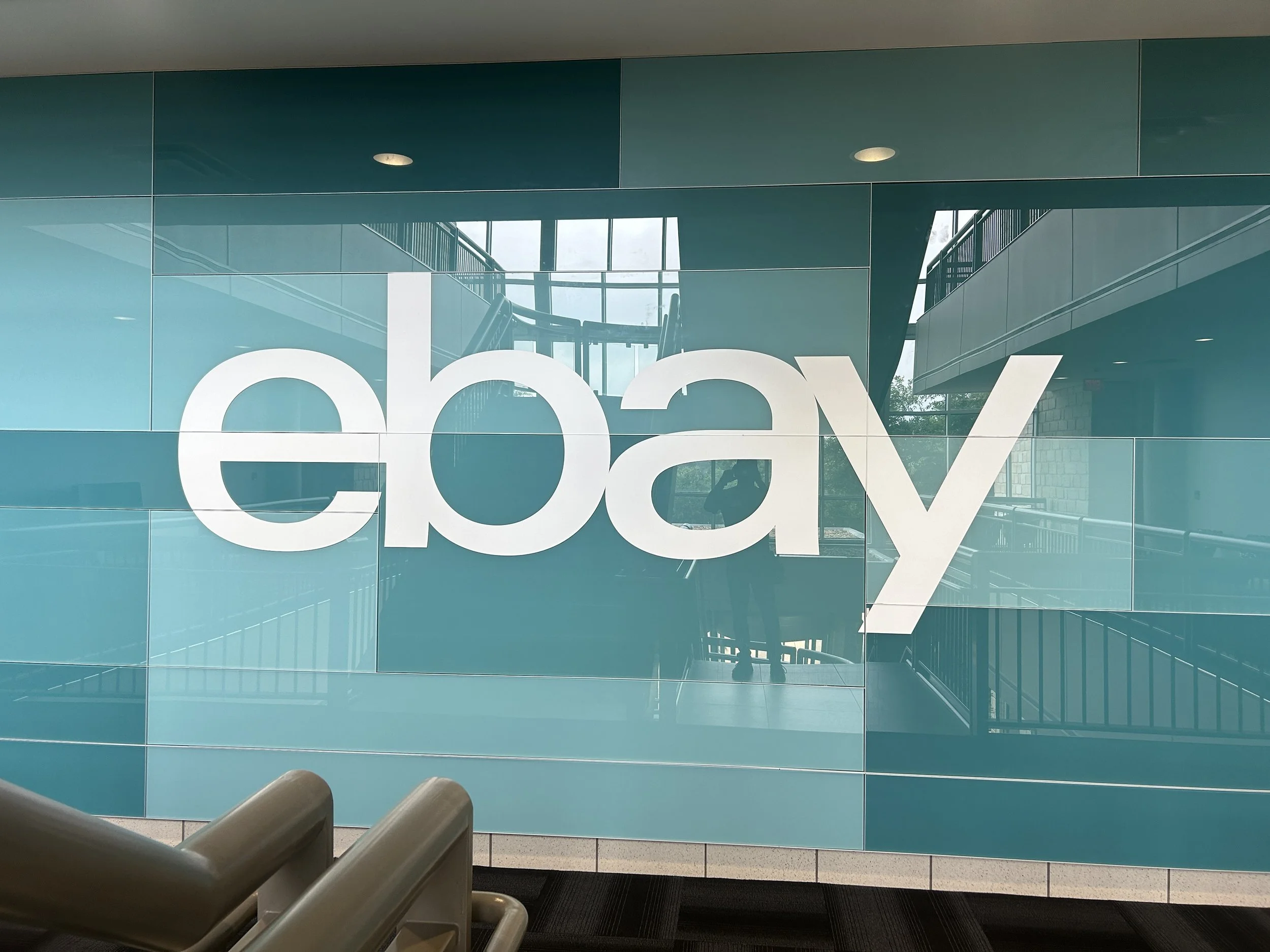 Installed eBay Austin wallcovering with layered teal environmental graphics behind glass, featuring large white logo in office lounge area.