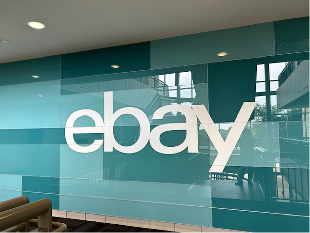 eBay Austin interior wallcovering installation with teal graphic panels behind glass, featuring large white logo and environmental branding in office lobby area.