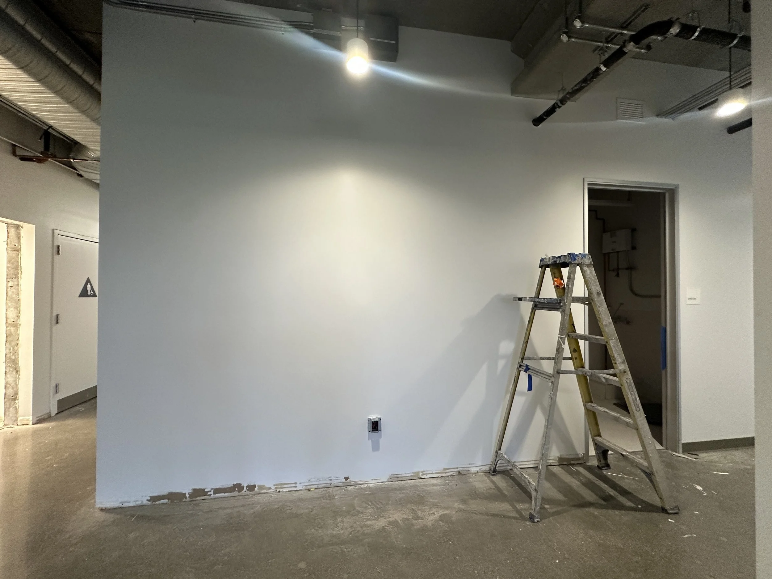 Office wall shown before installation of acoustic felt panels with ladder and open workspace during construction phase.