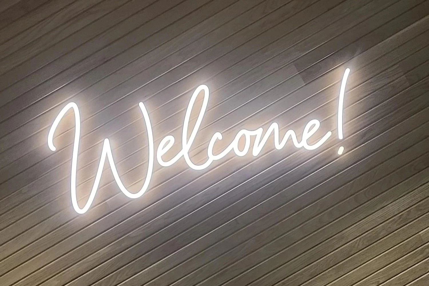 Installed eBay Austin faux neon “Welcome” sign mounted on wood slat wall, creating illuminated branded entry feature within office environment.