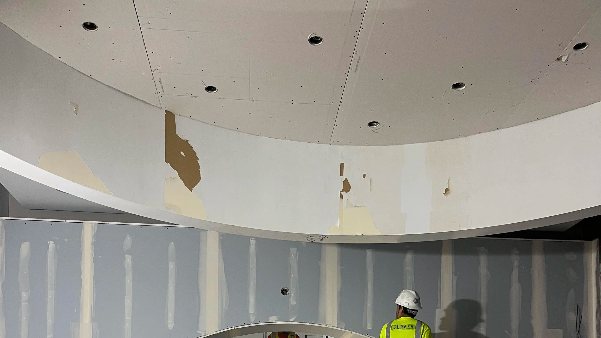 Curved drywall soffit under renovation with surface patching and preparation for finishing, featuring recessed lighting cutouts in a commercial interior construction project.