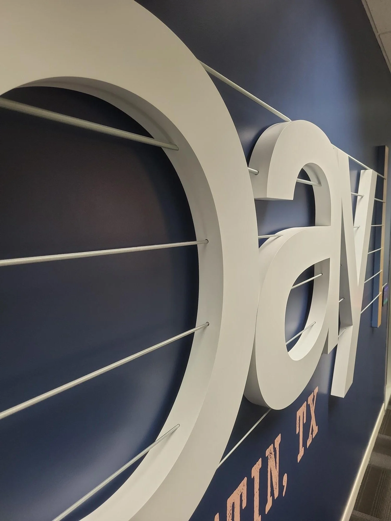 Side profile view of eBay Austin dimensional signage showing depth of raised logo letters and steel cable guitar string elements on navy feature wall.