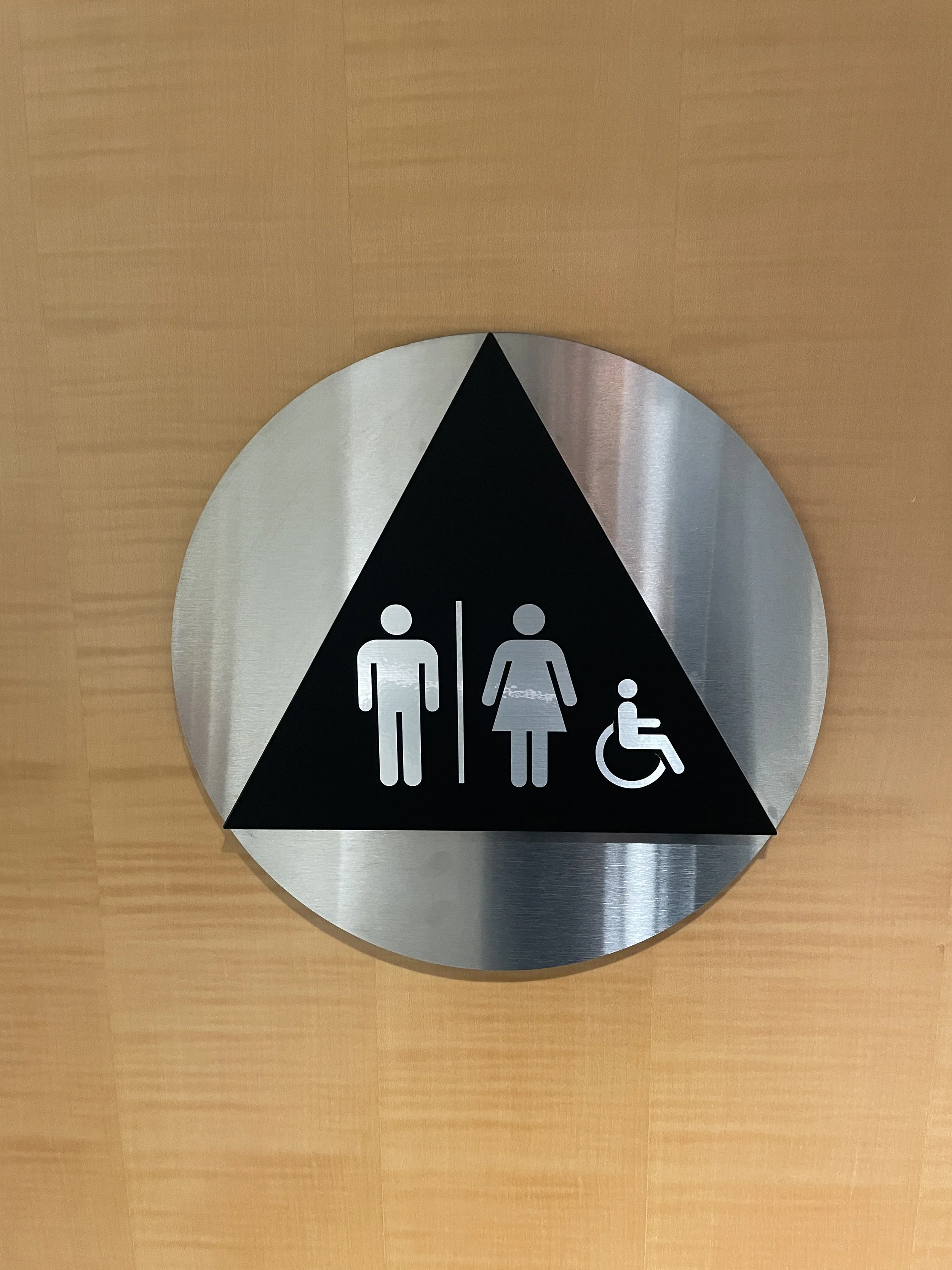 Brushed metal ADA restroom sign with male, female, and wheelchair accessibility symbols mounted on wood wall for compliant interior wayfinding.