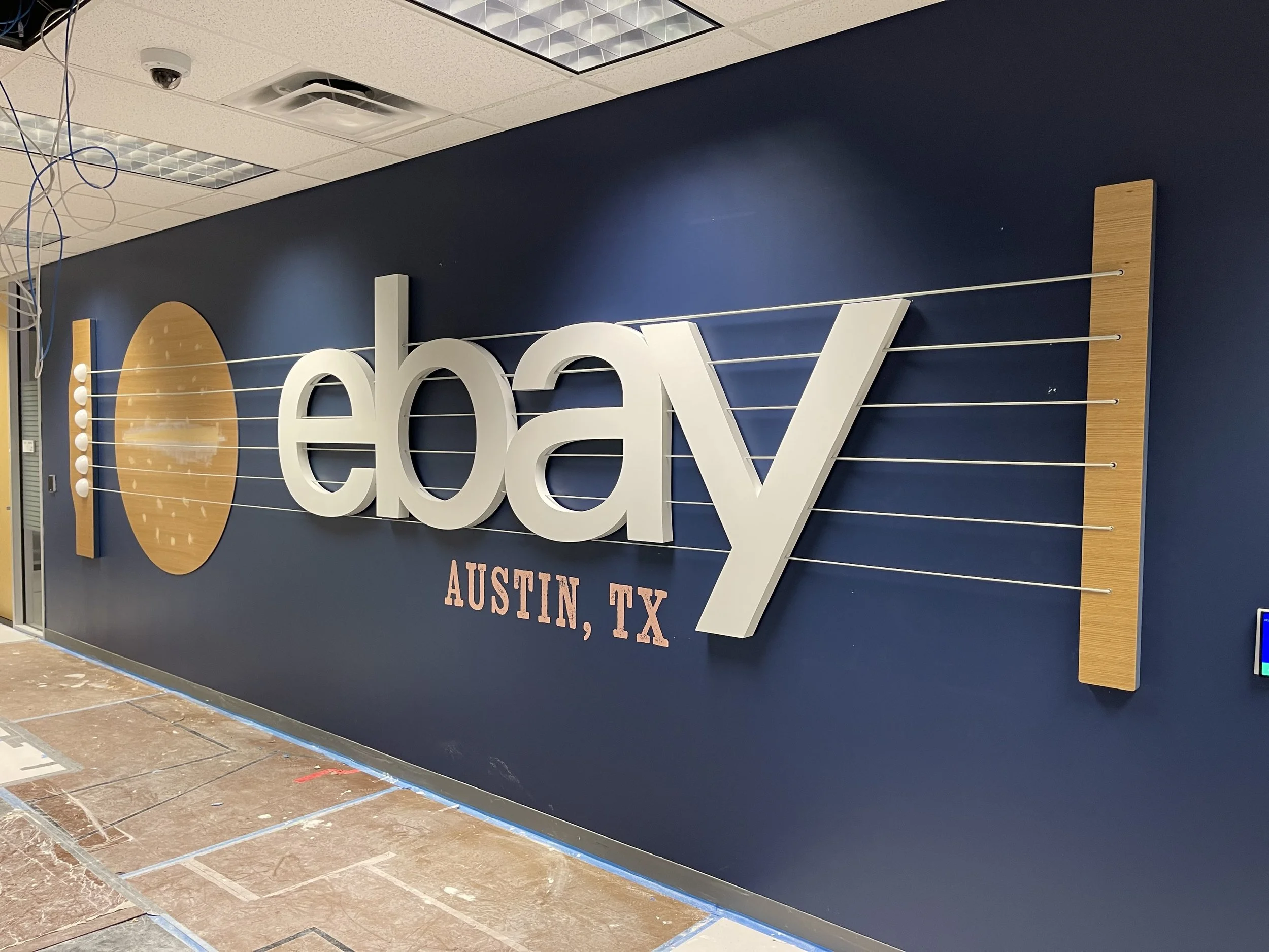 eBay Austin dimensional feature wall installation showing raised logo letters with white oak guitar-inspired elements and steel cable details on navy painted wall prior to Texas disc installation.