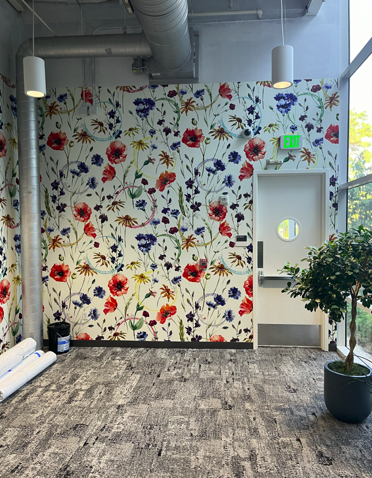 Installed floral wallcovering in amphitheatre hallway featuring colorful botanical pattern around a door with circular window and adjacent interior greenery.