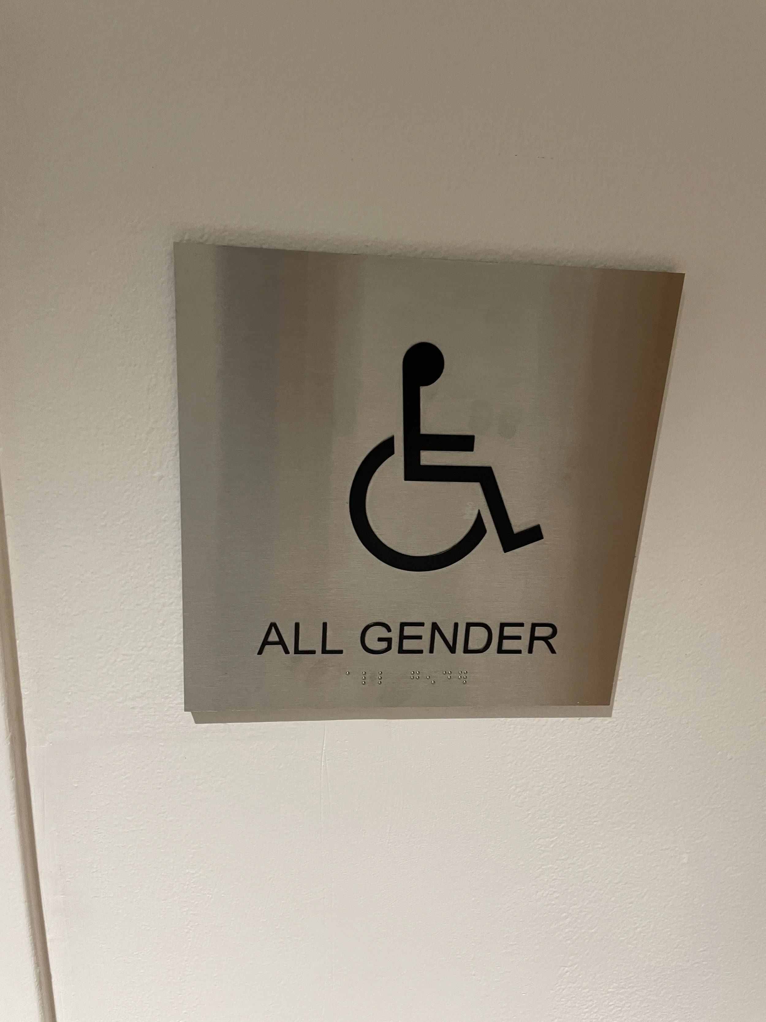 Brushed aluminum ADA all-gender restroom sign with wheelchair symbol and Braille mounted on interior wall for accessible wayfinding.