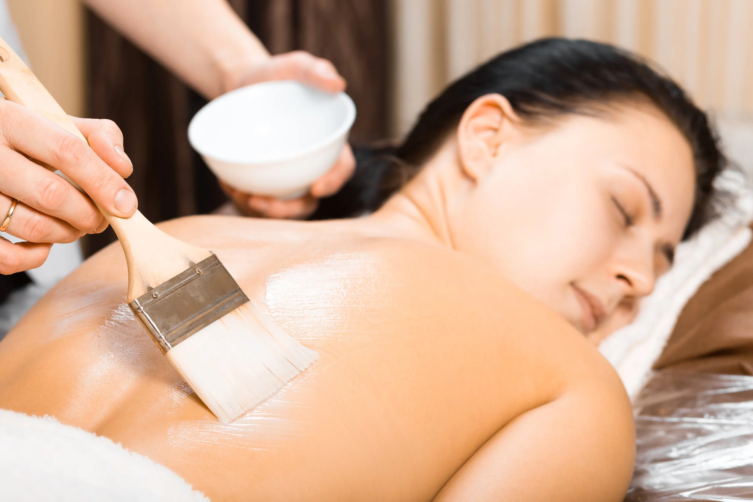 A woman receiving a massage with a brush being applied to her back with soap or oil, in a spa setting.