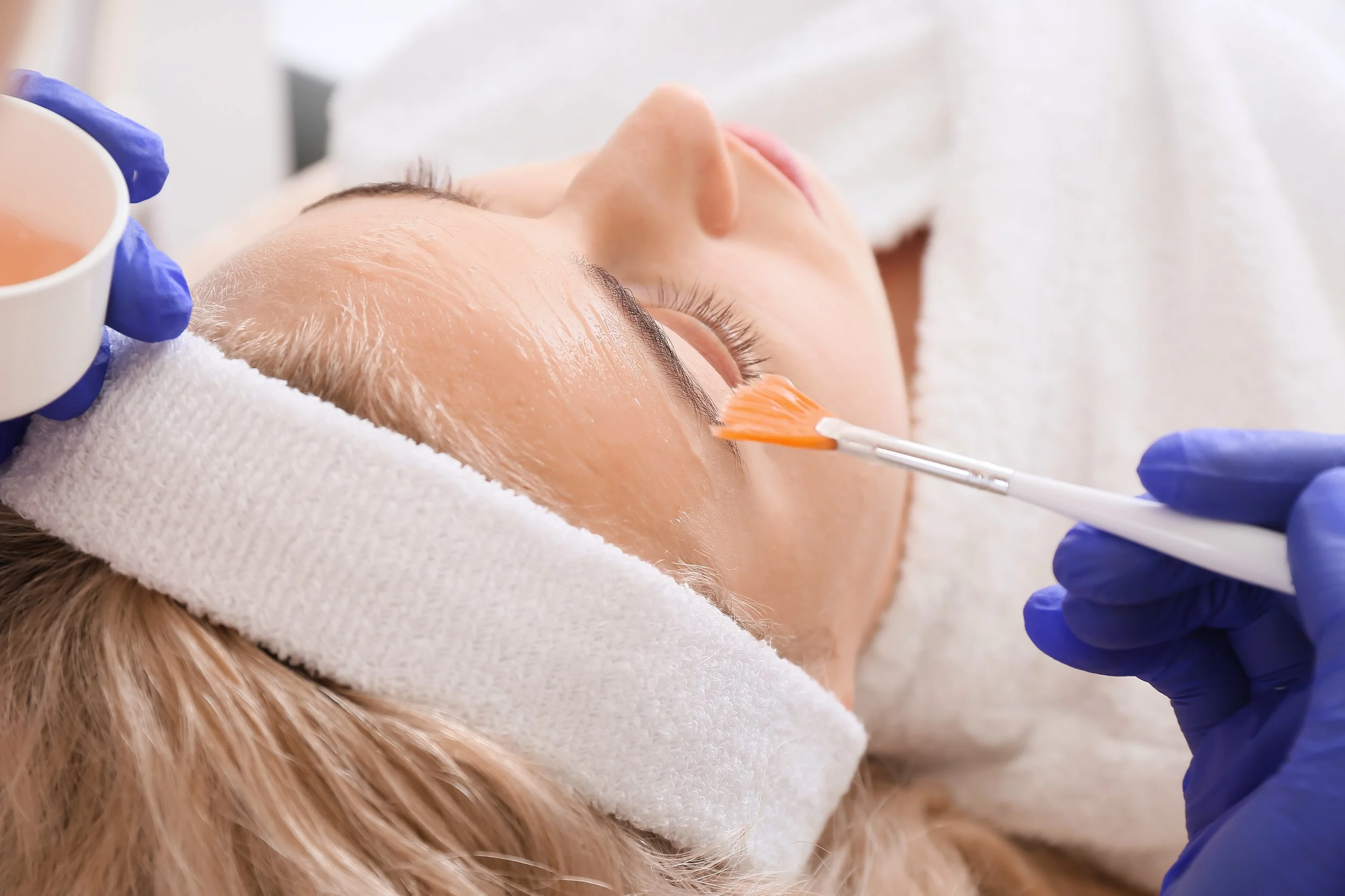 A woman receiving a facial treatment, lying down with her eyes closed, as a skincare professional applies a product with a brush on her face, wearing gloves.