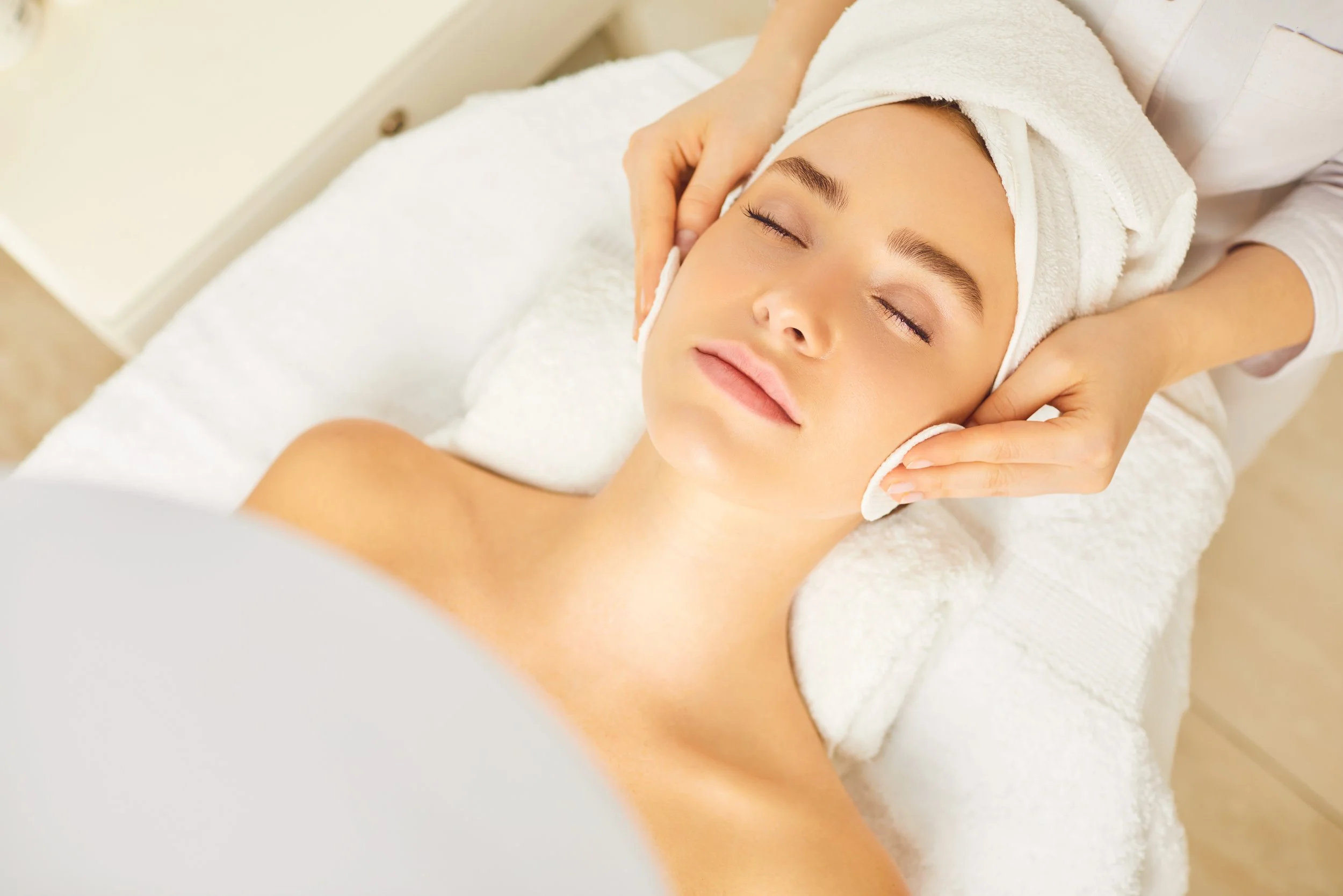 Woman receiving a facial massage at a spa, lying with eyes closed, towel wrapped around her head.