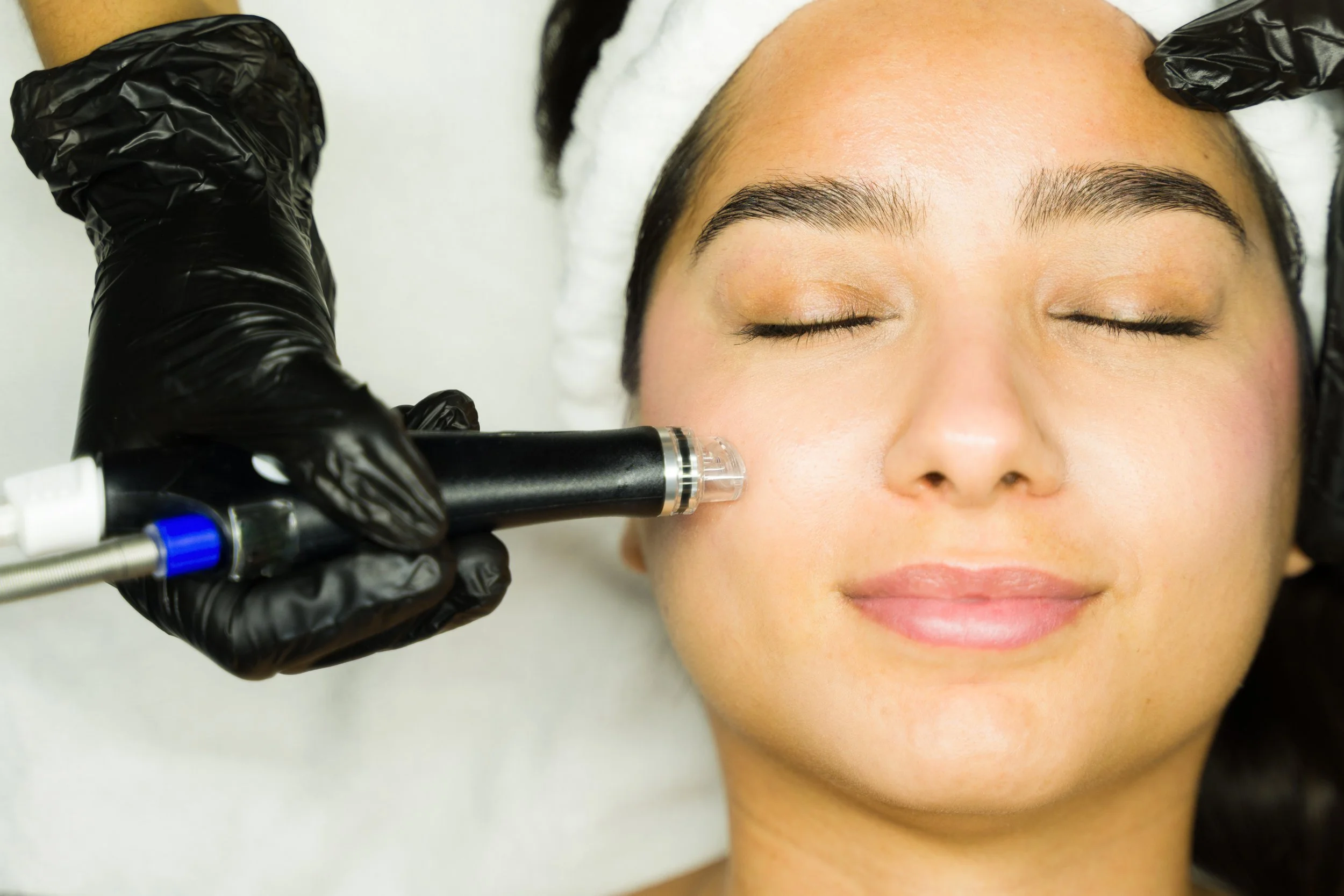 A woman with closed eyes relaxing during a facial treatment, while a practitioner uses a handheld device on her cheek.