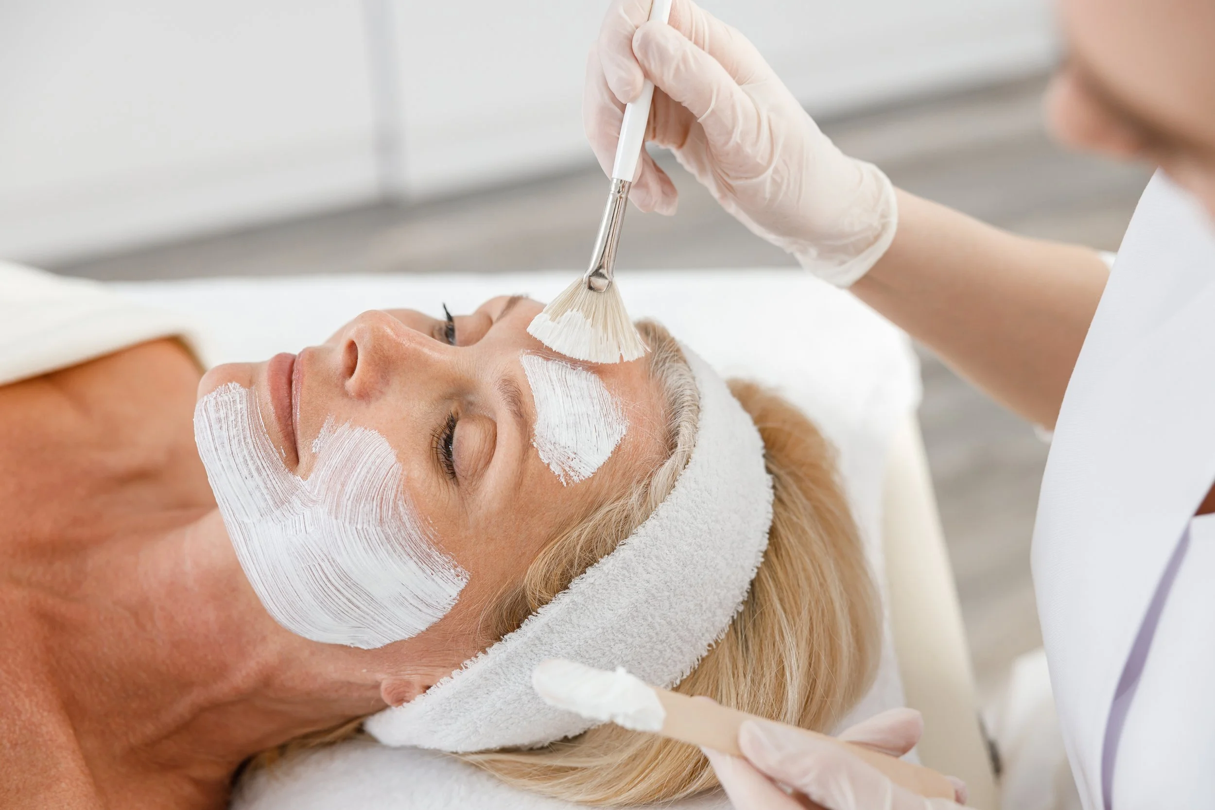 A woman receiving a facial treatment at a spa, with her eyes closed, lying down with a white towel wrapped around her head, while a skincare professional applies a white facial mask with brushes.