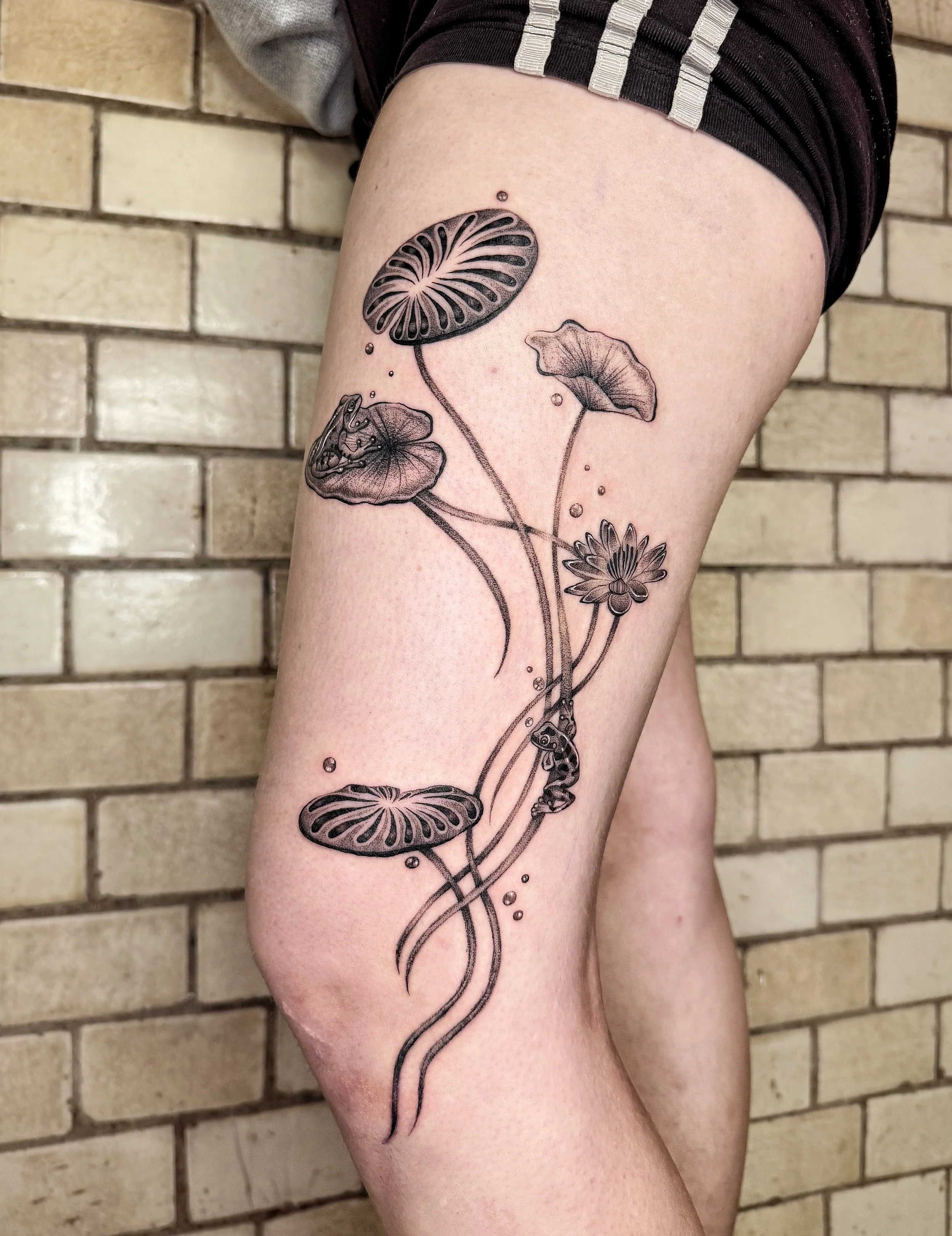 Schwarze Tätowierung eines Wasserpflanzenmusters auf dem Oberschenkel einer Person, mit Blättern, Stängeln, kleinen Blüten und einer Muschel, vor einer Ziegelwand.