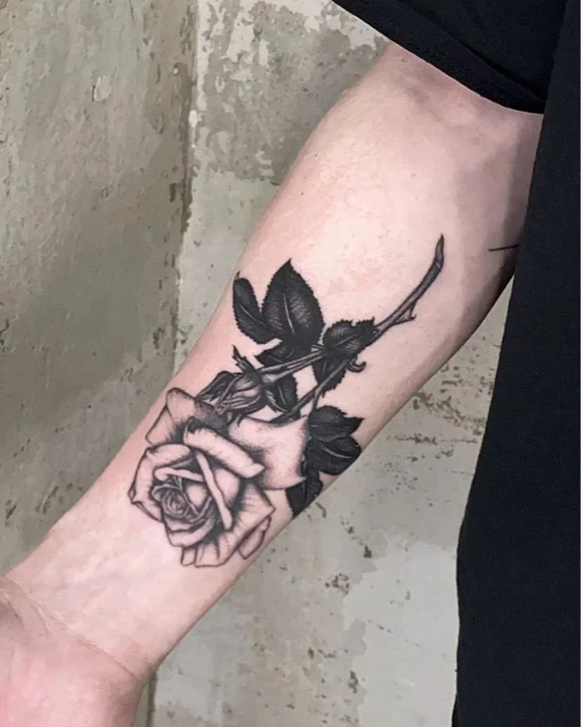 Schwarze Tätowierung einer Person, die eine Rose in einer Hand hält, mit einigen Blättern und einer Fliege auf der Rose, auf dem Unterarm.