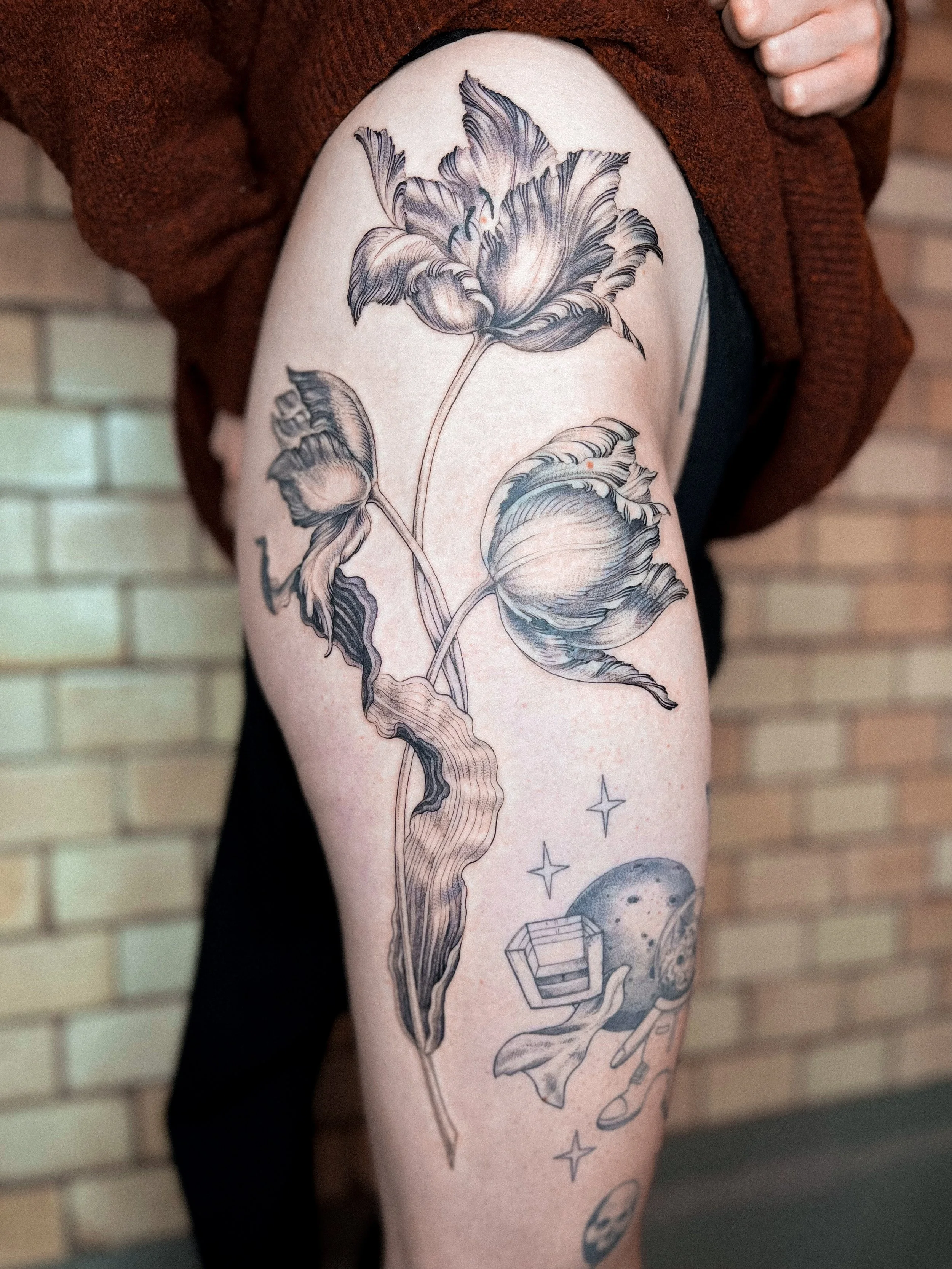 Schwarz-weiße Tätowierung einer großen Blume mit Blättern auf einem Oberschenkel.