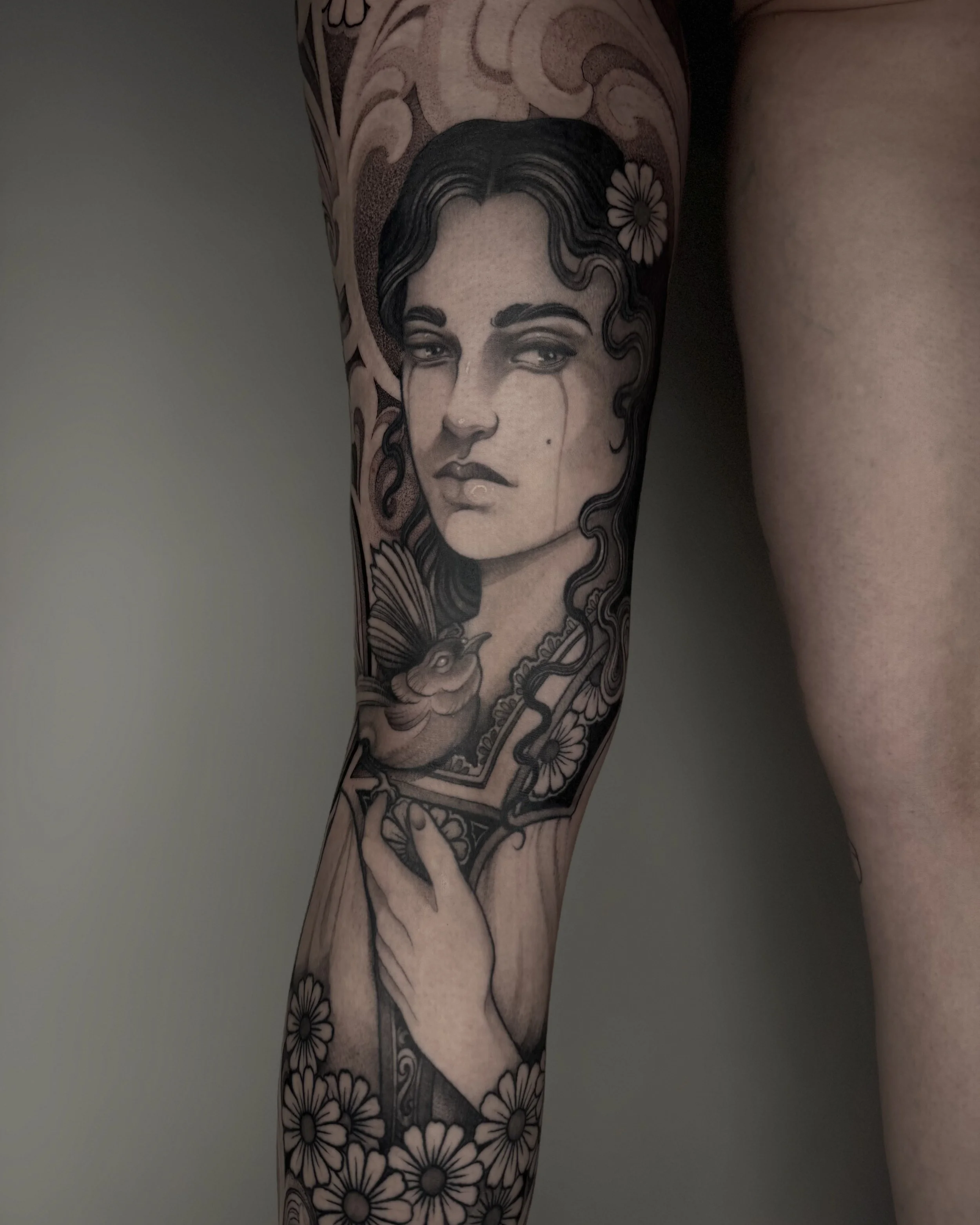 Tattoo eines Porträt eines Mädchens mit dunklen, gewellten Haaren, Blumen, einem Vogel und einer Hand, die eine Spiegelscheibe hält.