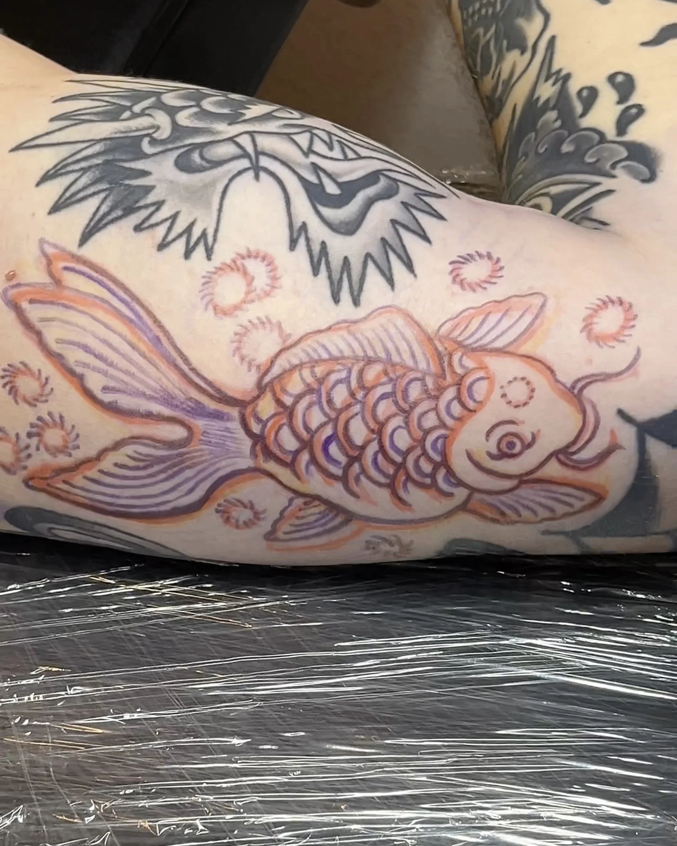 Ein farbenfrohes Tattoo eines Goldfisches und eines Drachen auf einem Arm, mit einem schwarzen Hintergrund.
