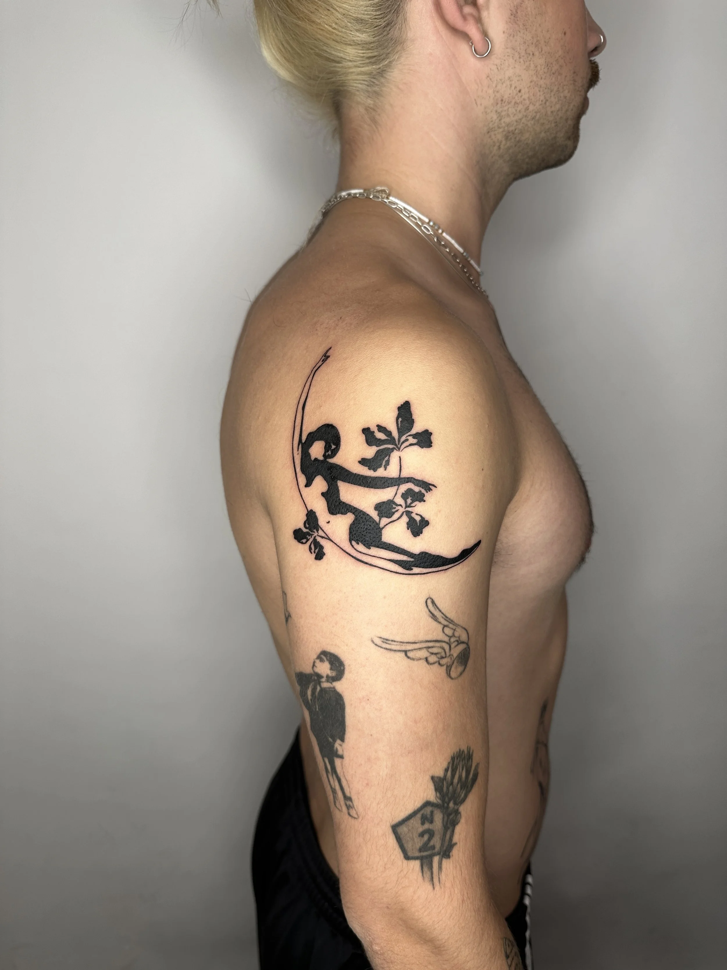 Ein Mann mit Tattoos auf dem linken Oberarm und der Schulter, einschließlich eines Männchens auf einer Schaukel, eines Mädchens, eines Flügels, eines Hauses mit der Zahl 22 und einer Blume.