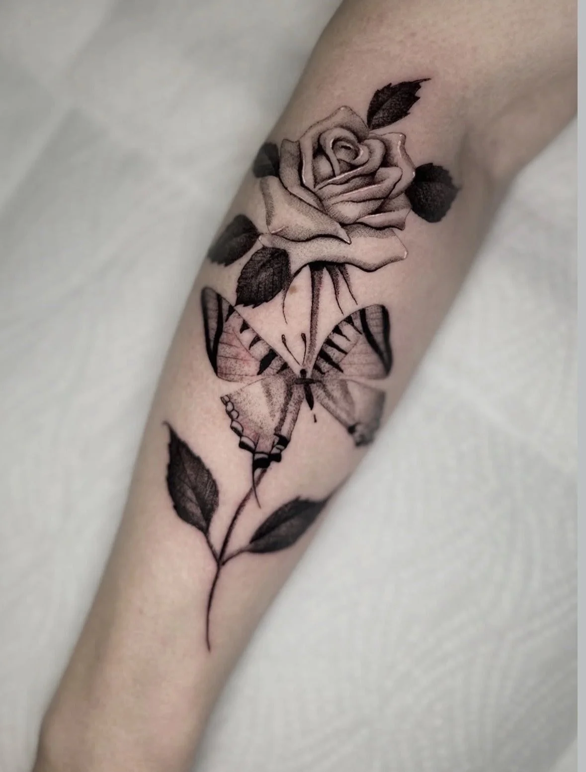 Tattoo eines stilisierten Rosenstocks mit einer rosa Blume und einer Schmetterlingsillustration auf dem Unterarm.