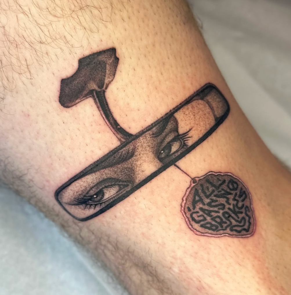 Tattoo eines Mannes auf seinem Arm. Es zeigt eine Lupe mit einem menschlichen Gesicht, insbesondere Augen und Nase. An der Spitze der Lupe hängt ein Seelensymbol mit dem Schriftzug 'ALL LOVE BEGINS'.