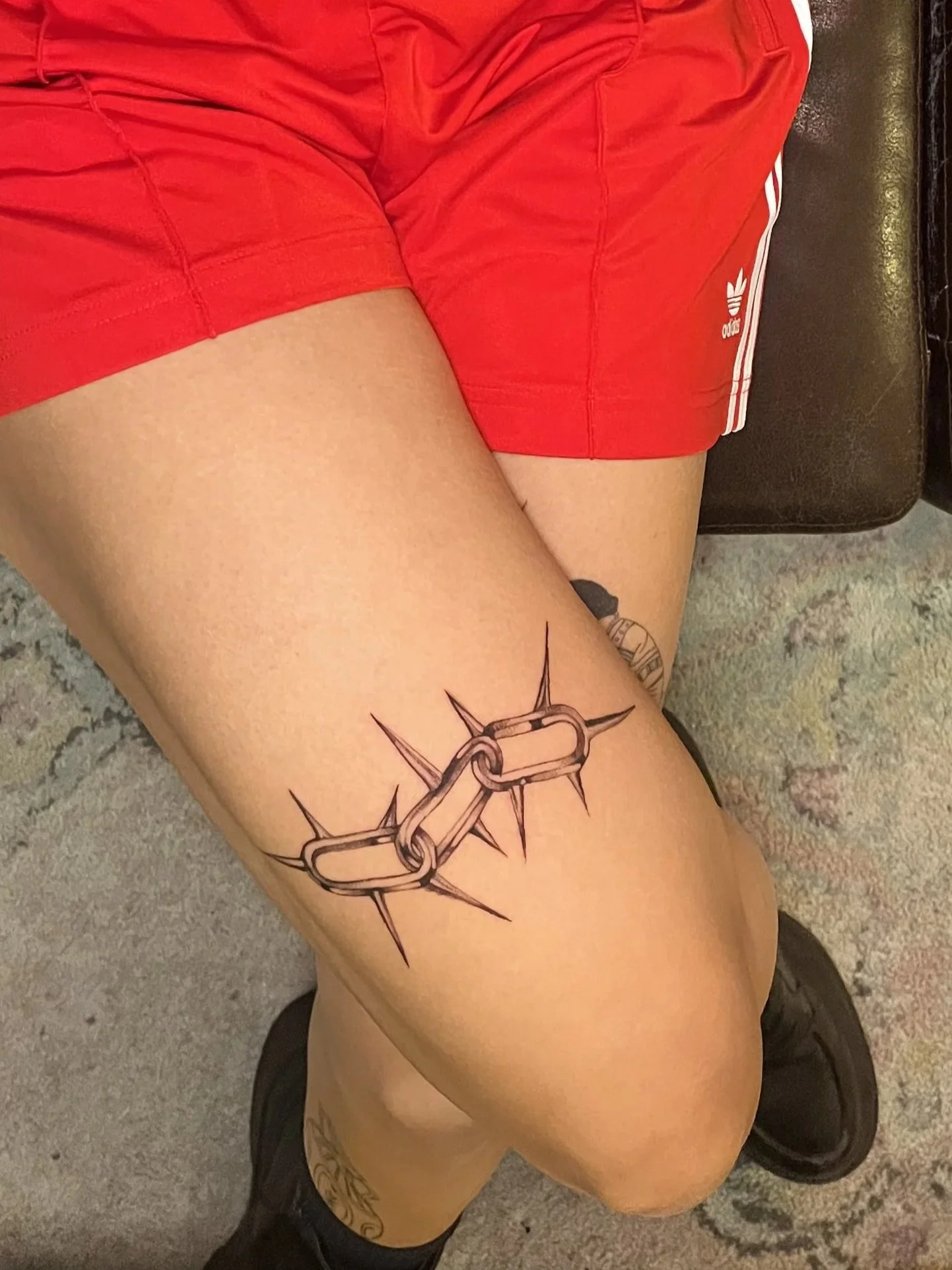 Tattoo eines Kettenglieds mit Dornen auf Oberschenkel einer Person, die rote Sportshorts trägt und auf einem Teppich liegt.
