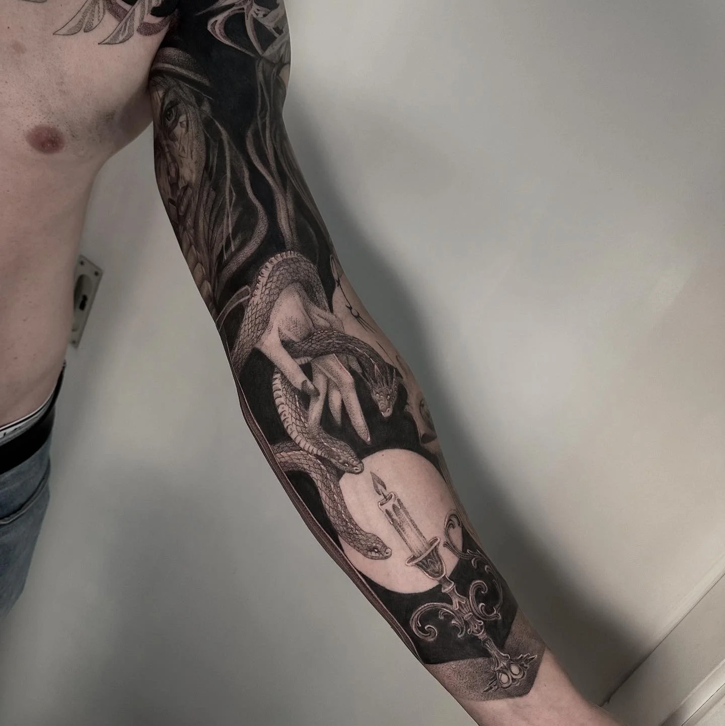 Schwarze Tätowierung eines Drachen, einer Kerze, eines Mondes und eines mystischen Motivs auf dem Arm eines Mannes.