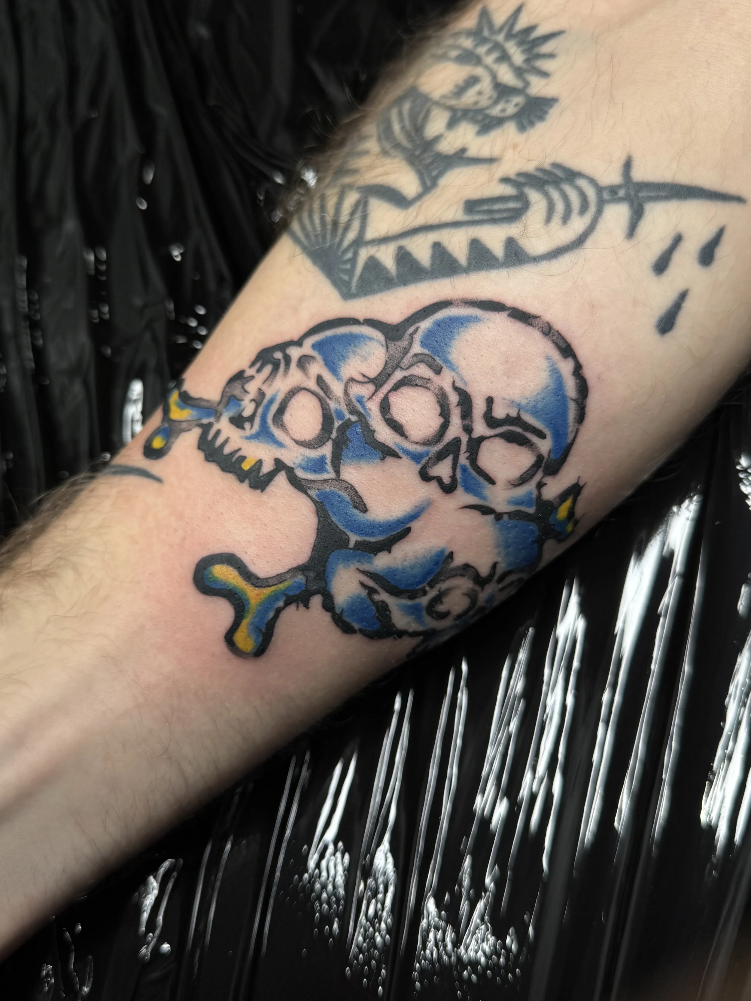 Tattoo eines grünen Skulls mit blauen und gelben Akzenten auf dem Unterarm.