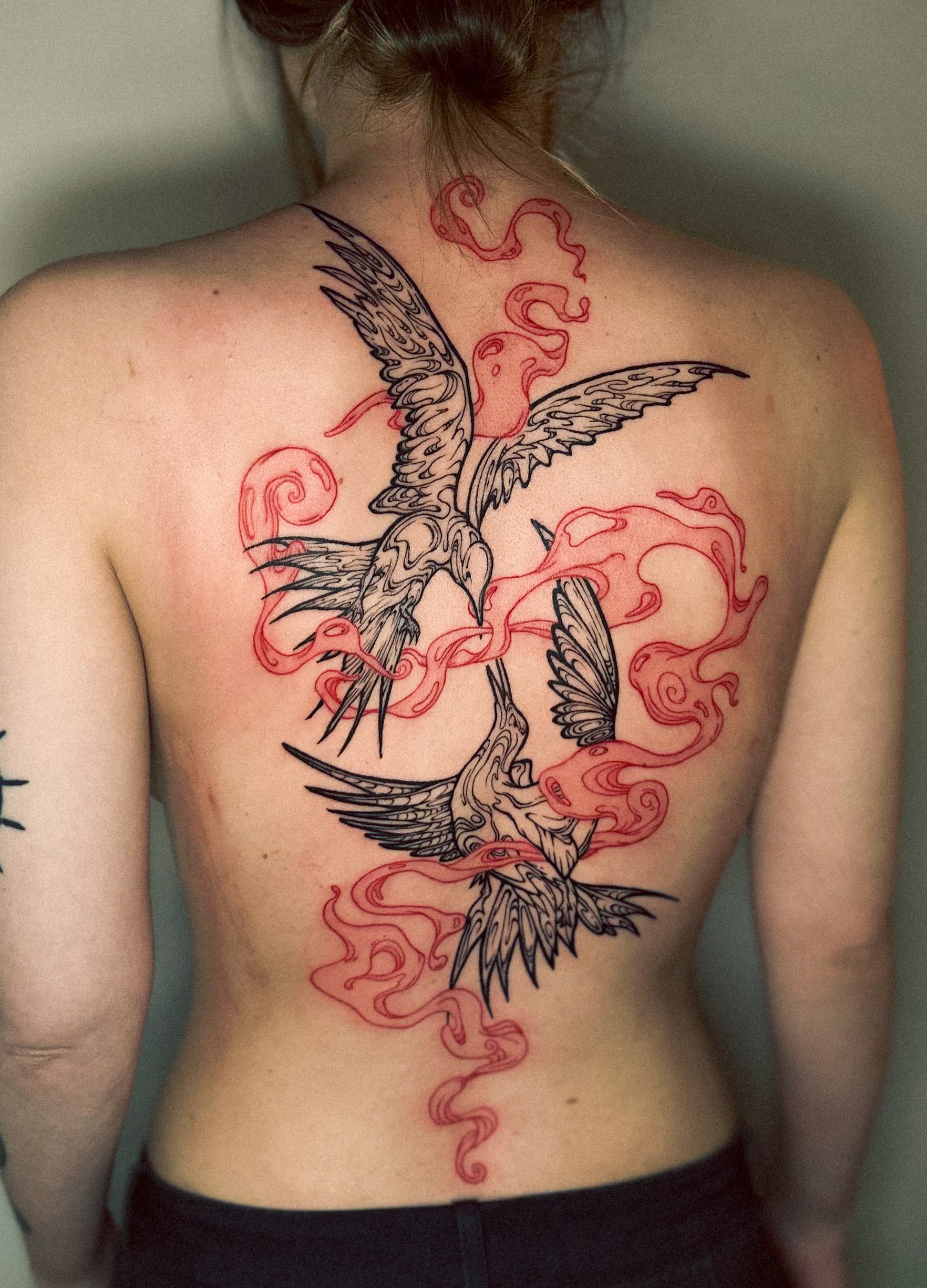 Ein Rücken mit einem großen Tattoo von zwei fliegenden Vögeln, umgeben von roten Rauch- oder Flammenmustern, in Schwarz und Rot.