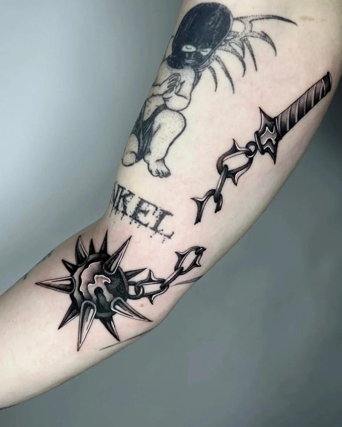 Schwarze Tätowierungen auf einem Arm, darunter ein Käfer, ein stilisiertes Schwert, ein Kreuz mit einer Kette, das Wort 'KIEL' und ein Stern mit einem Anker.