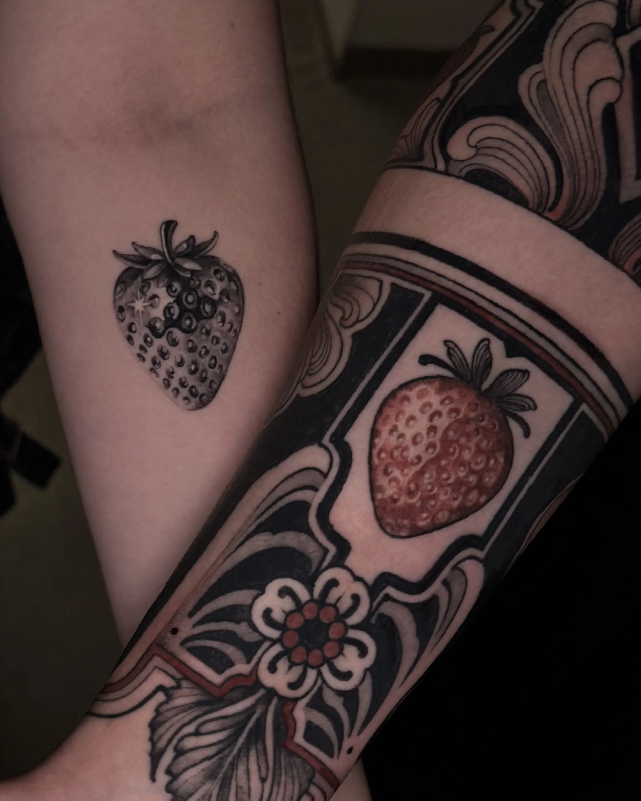 Tattoo eines Erdbeers auf dem Unterarm und ein größeres, detailliertes Erdbeer-Tattoo auf dem Oberarm eines Menschen, umgeben von dekorativen Mustern.